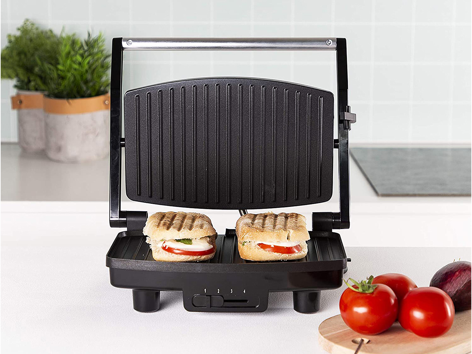 Schwarzer Panini-Grill mit zwei Sandwiches. Tomaten und Basilikum daneben. Weiße Küchenarbeitsplatte.