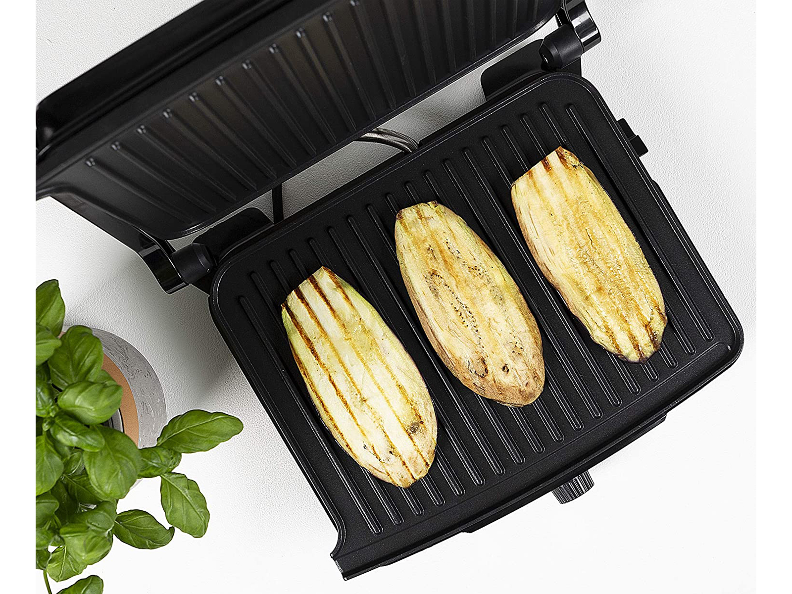 Gegrillte Auberginenscheiben auf einem schwarzen Grill. Grüne Pflanze im Hintergrund.