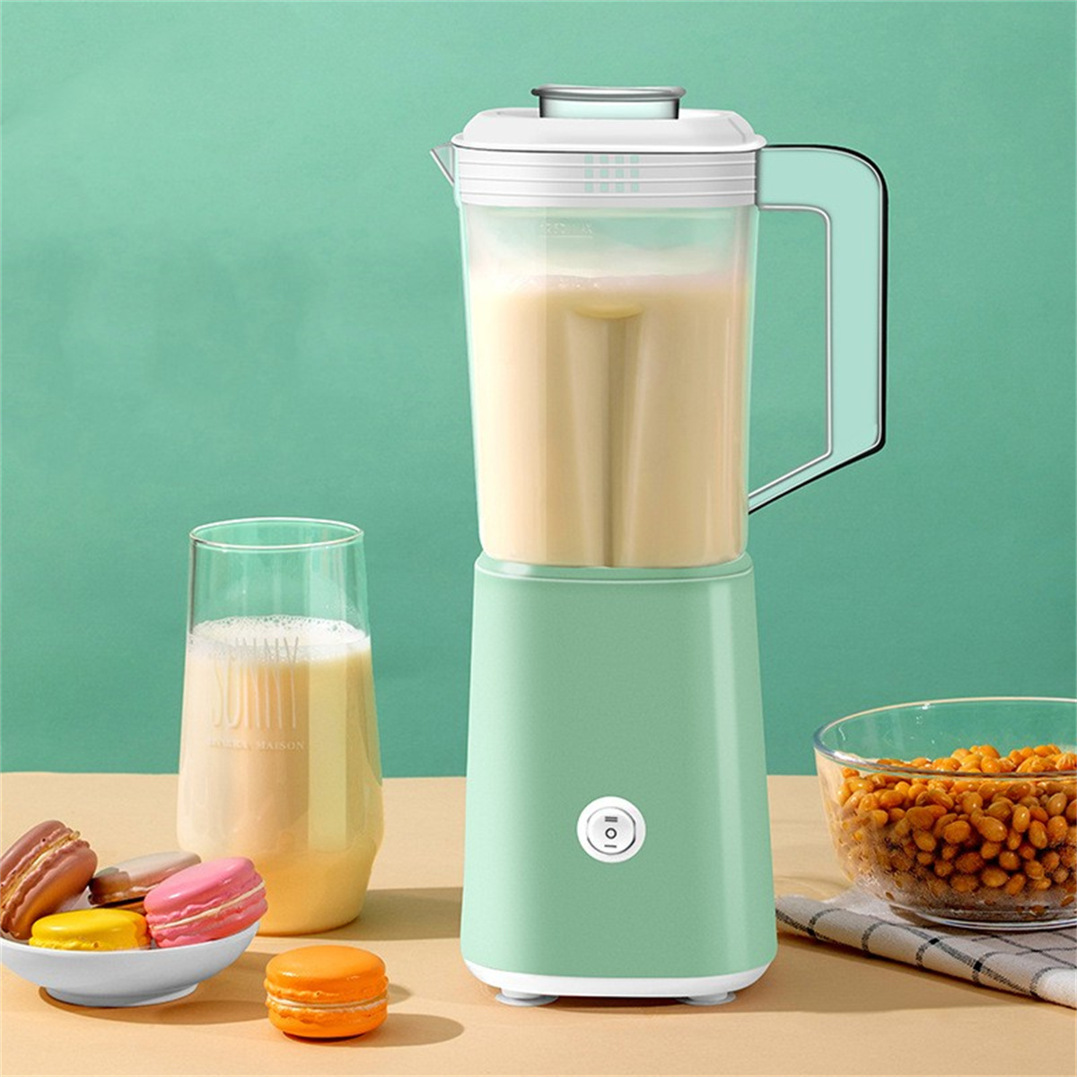 Mixer mit hellgrüner Basis und durchsichtigem Behälter, gefüllt mit hellbeiger Flüssigkeit. Neben Macarons.