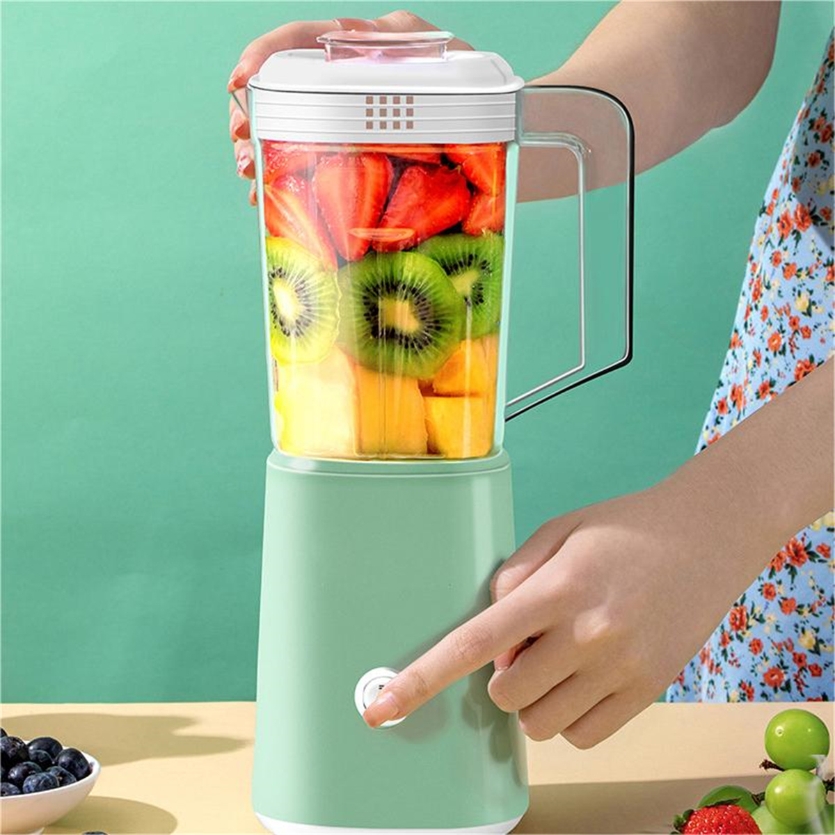 Eine Hand hält einen Mixer mit geschnittenem Obst. Eine Person drückt den Einschaltknopf.