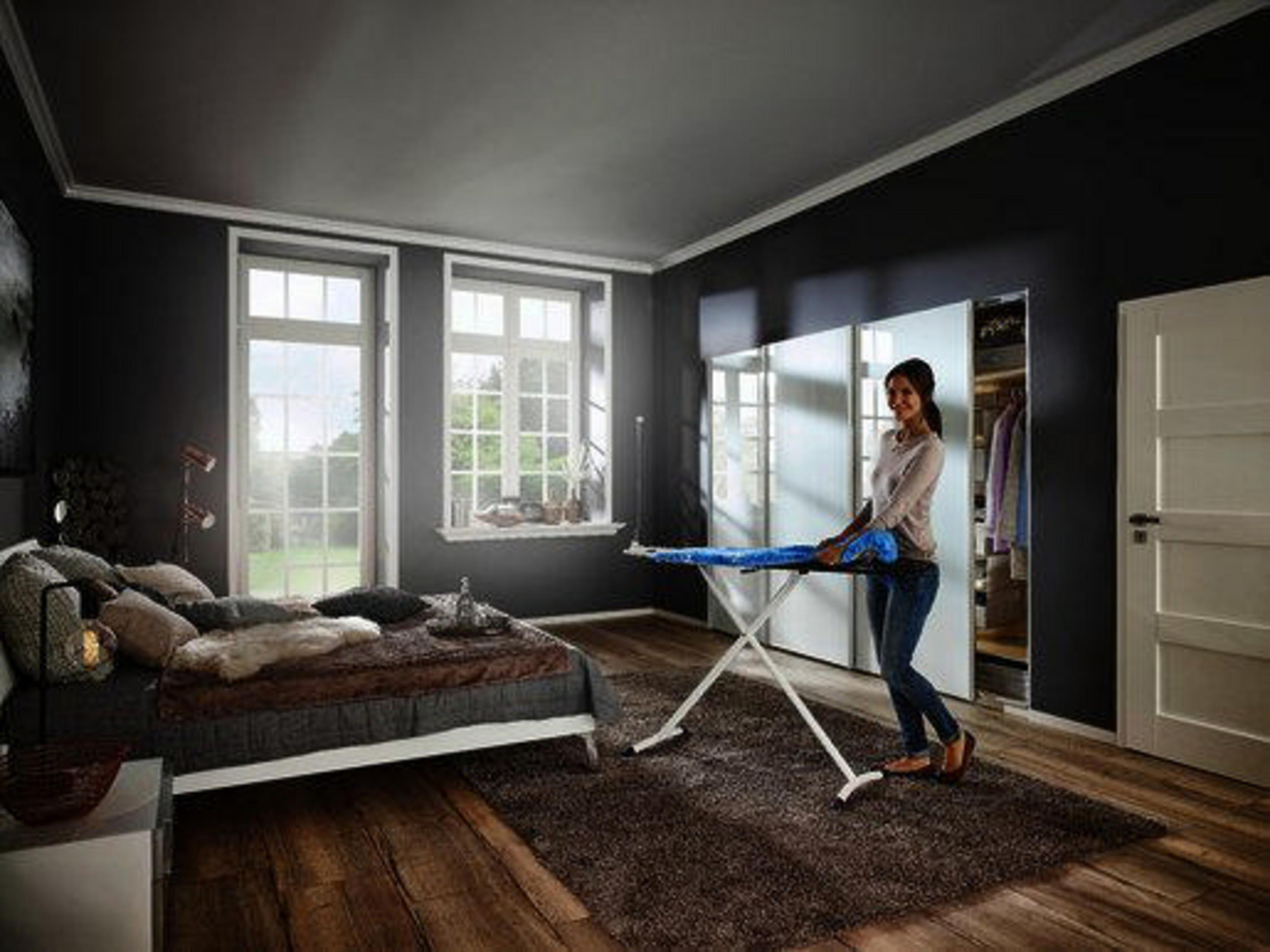 Kobieta prasuje ubrania w sypialni. Jest łóżko, okna i ciemne ściany.