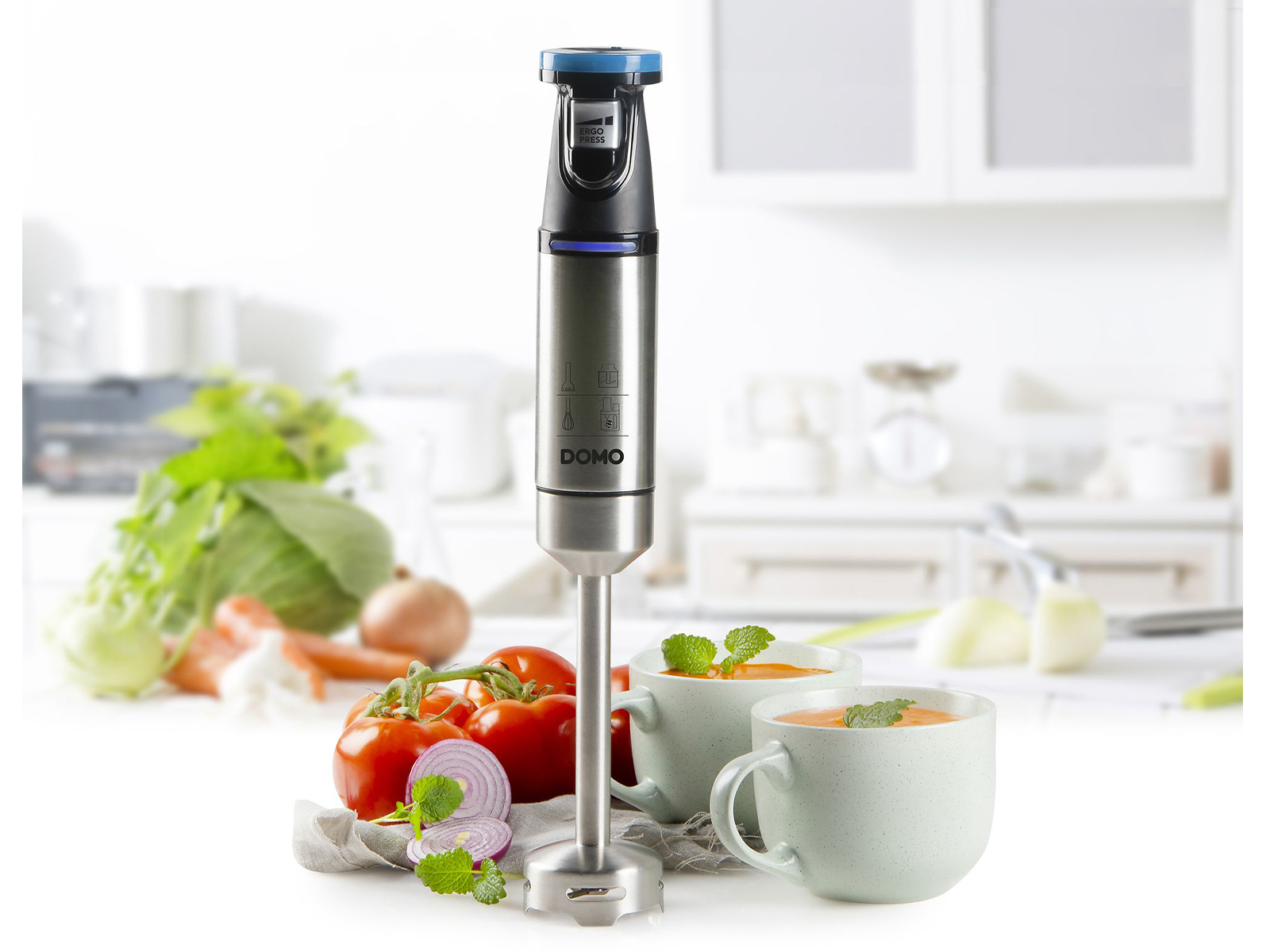 Ein silberner Stabmixer mit Gemüse und zwei Tassen Suppe in einer Küchenumgebung.