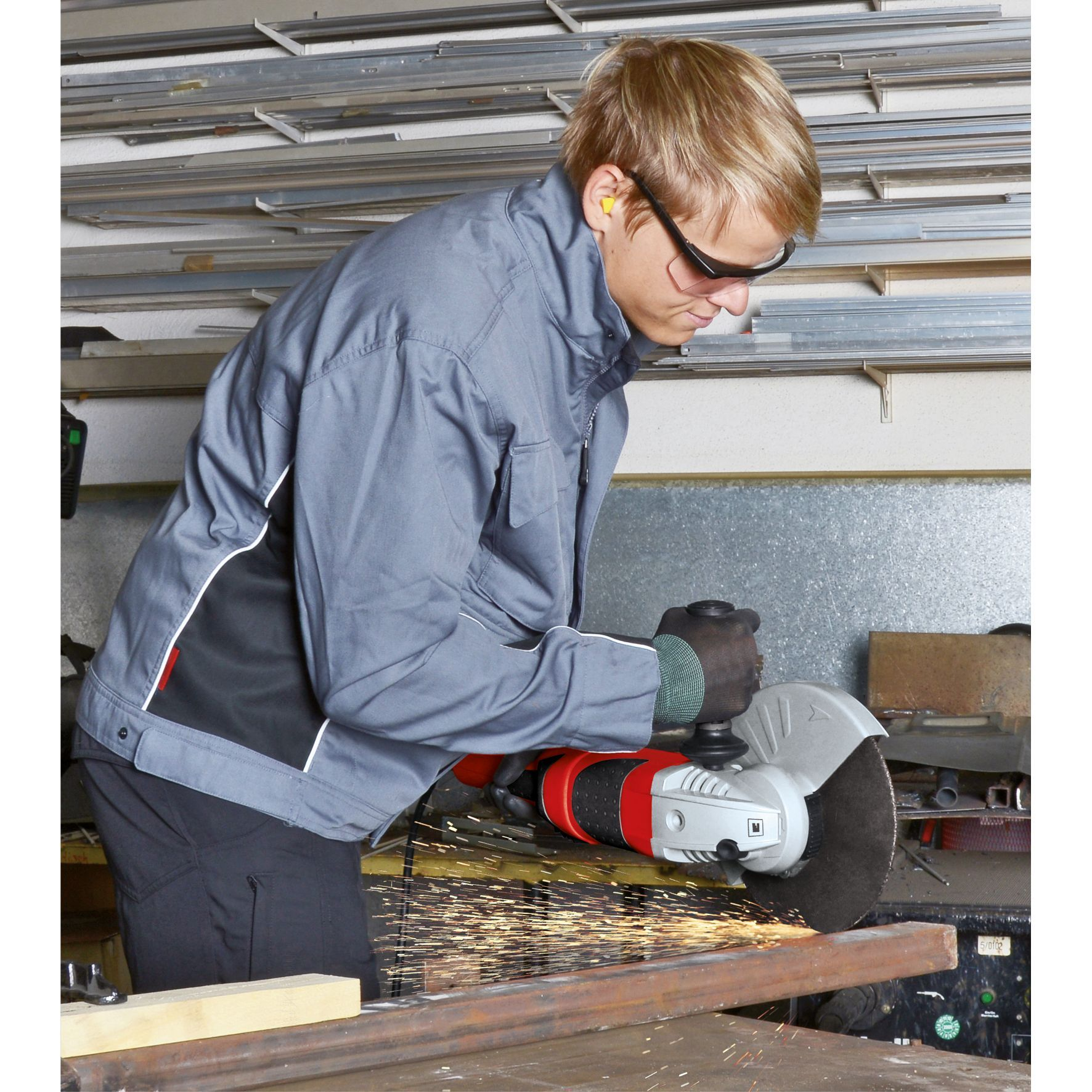 Mężczyzna używający szlifierki kątowej, w okularach ochronnych, zatyczkach do uszu i rękawicach. Iskry lecą z metalu.
