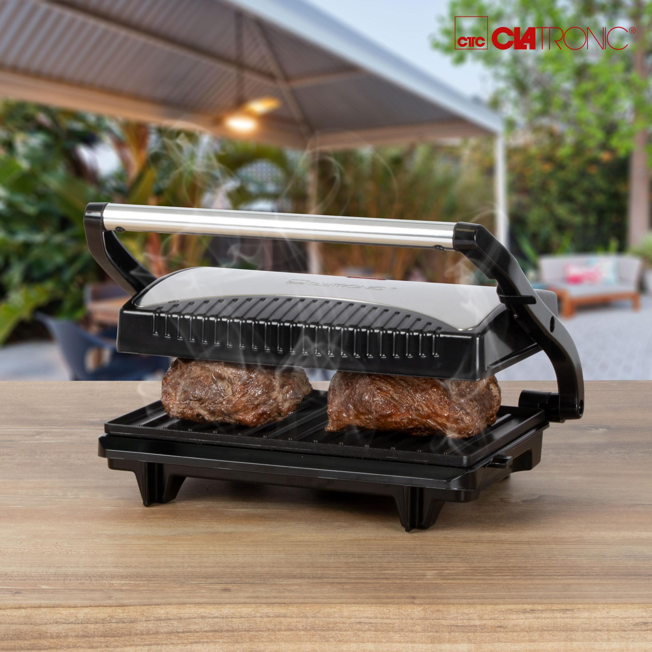 Ein Indoor-Grillgerät mit zwei gebratenen Steaks auf einem Holztisch, Rauch steigt auf.