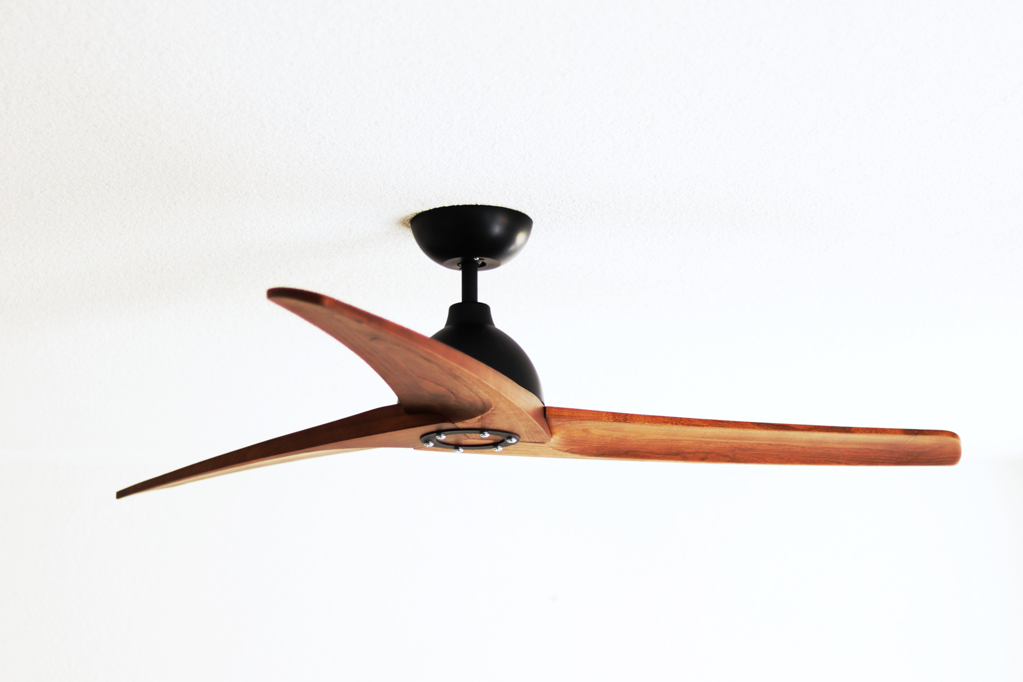 Deckenventilator mit drei Holzflügeln und einem schwarzen Motor. Er ist an einer weißen Decke montiert.