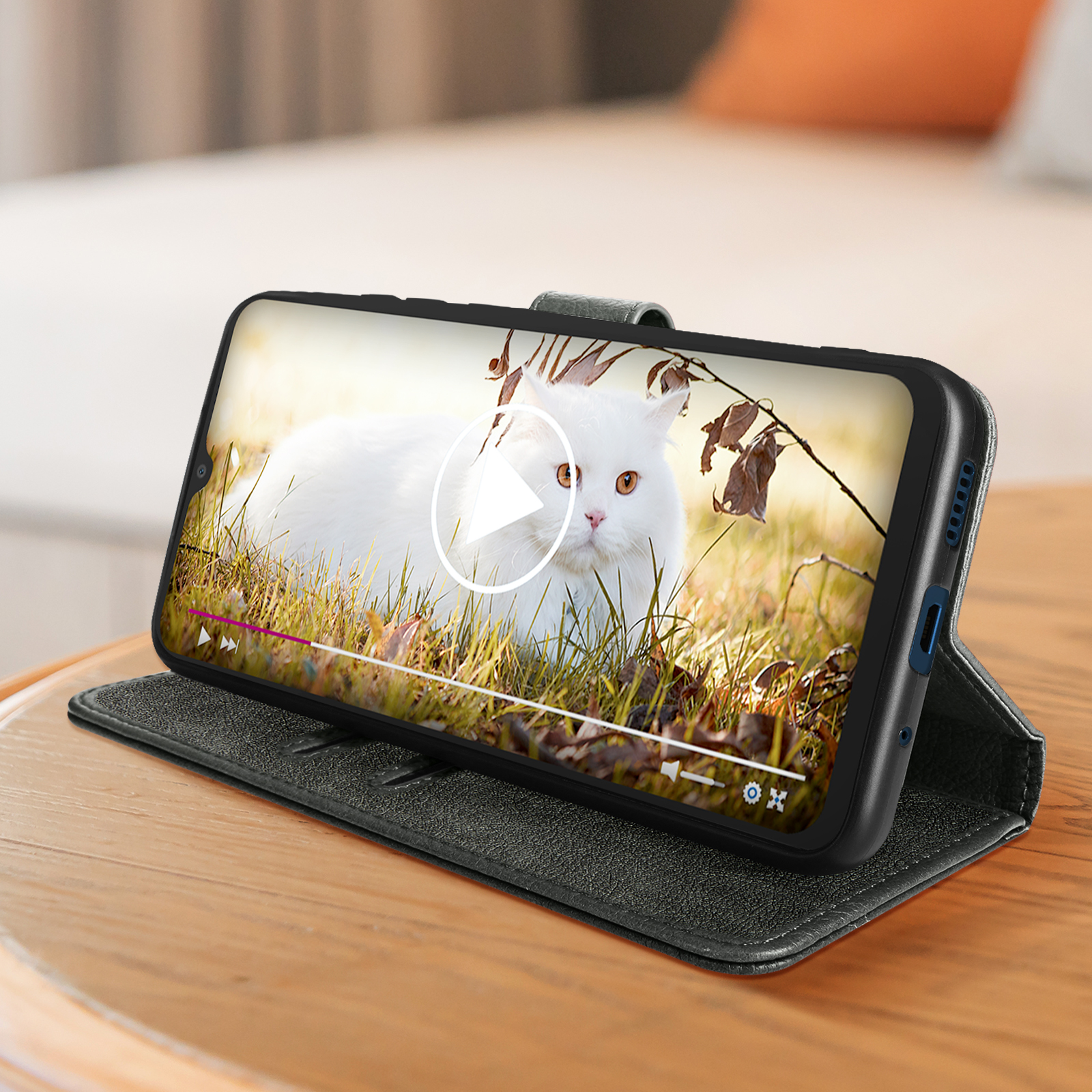 Schwarzes Telefon in dunkelgrüner Hülle, zeigt Video einer weißen Katze auf Holztisch.