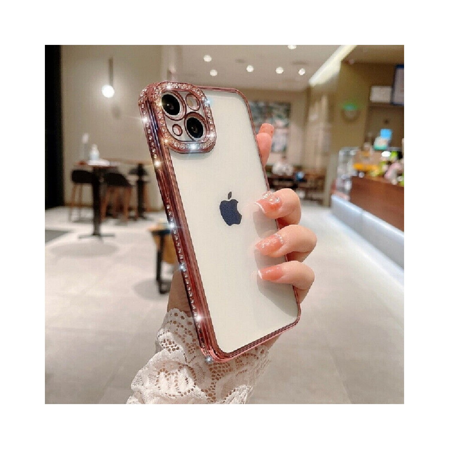 Una persona che tiene in mano un iPhone bianco con una custodia oro rosa con strass. Sfondo del caffè.