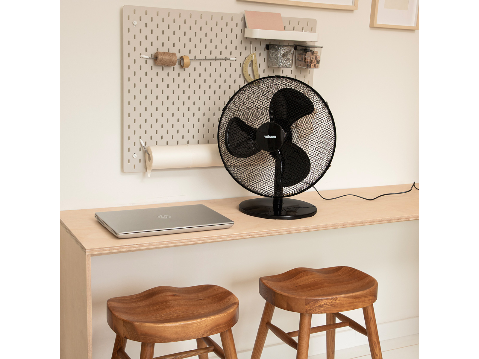 Ein schwarzer Ventilator auf einem Schreibtisch mit Laptop, Hockern und einer Lochwand im Hintergrund.