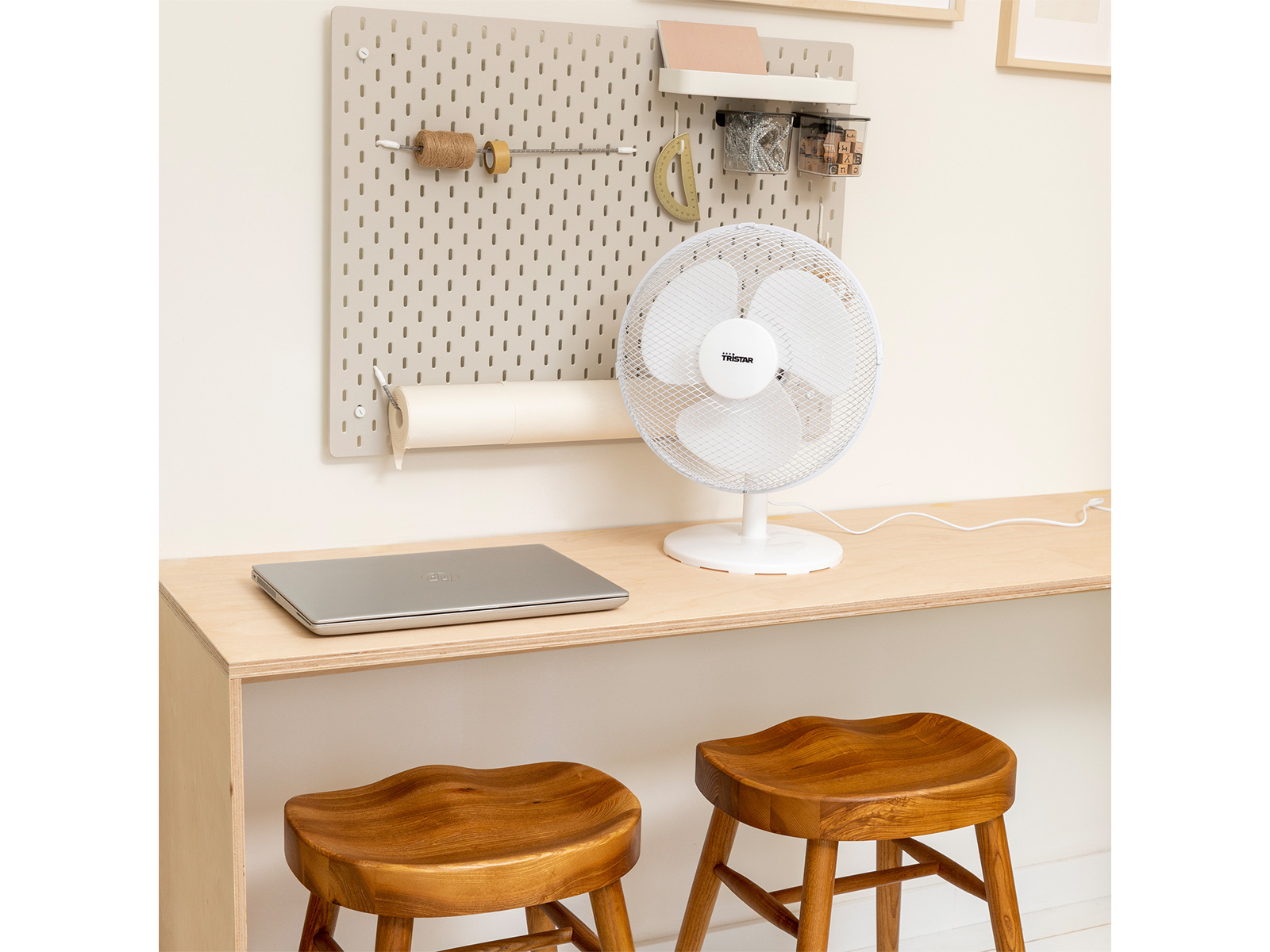 Schreibtisch mit Ventilator, Laptop und Lochwand an der Wand. Zwei Holzschemel. Weißer Hintergrund.