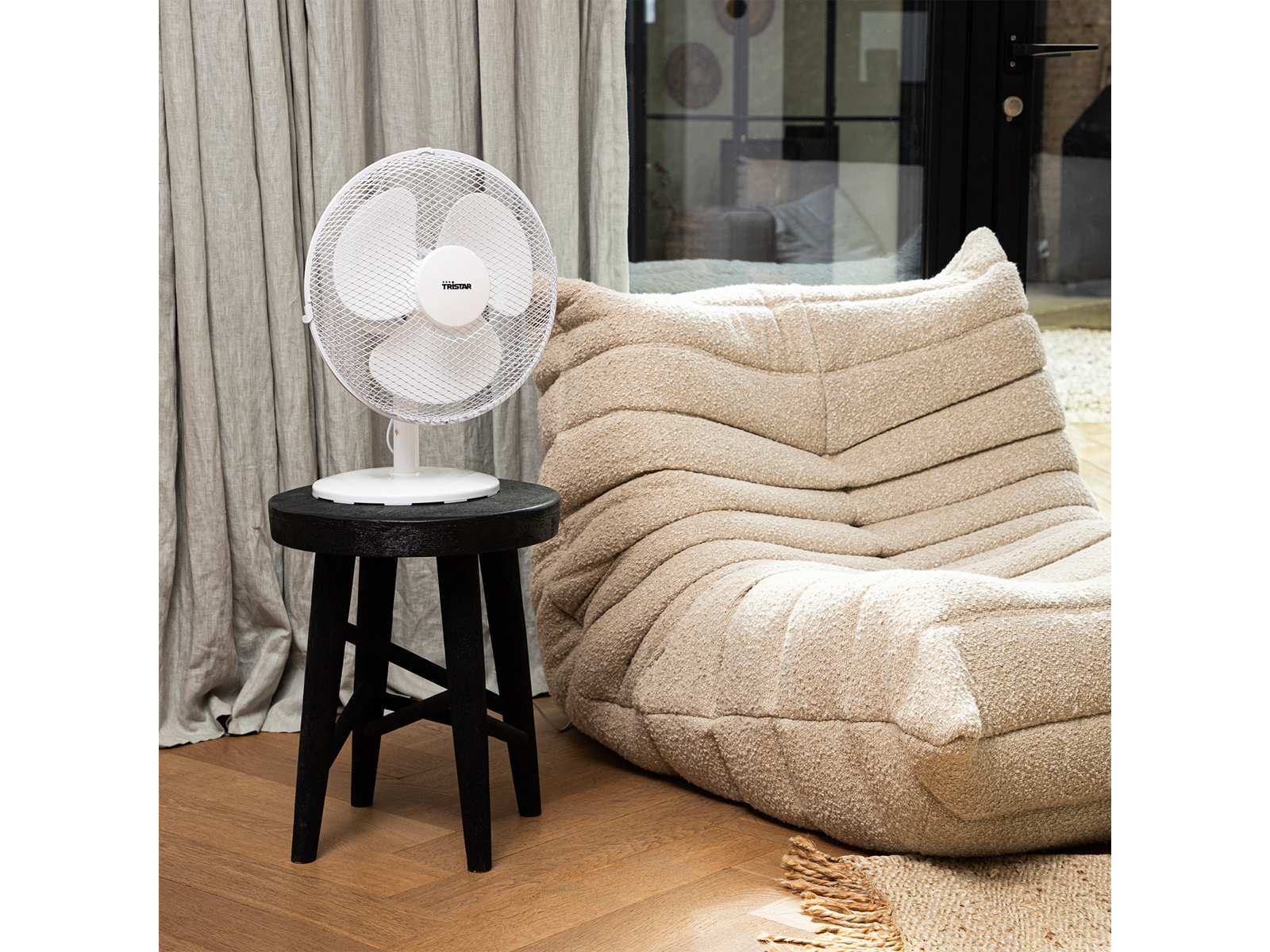 Ein weißer Ventilator auf einem schwarzen Hocker und eine beige, strukturierte Couch.