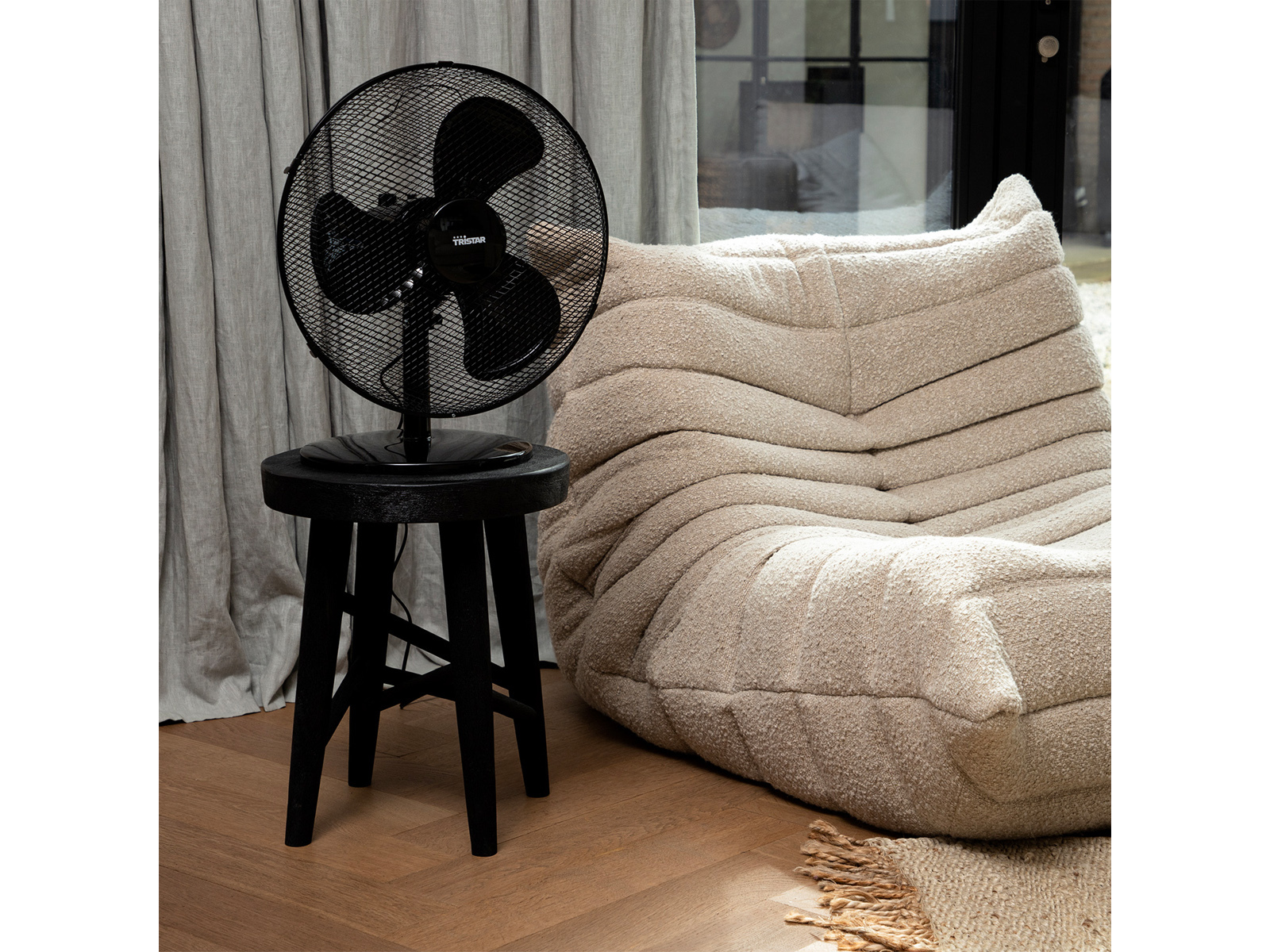 Ein schwarzer Ventilator auf einem schwarzen Hocker, neben einem beigefarbenen Sofa und einem Teppich.
