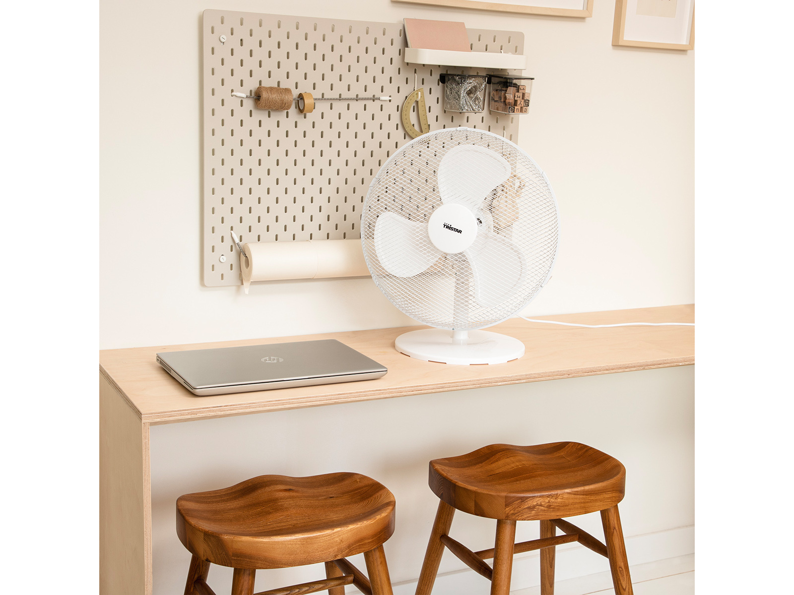 Schreibtisch mit Laptop, Ventilator und Lochwand. Zwei Holzstühle im Vordergrund.