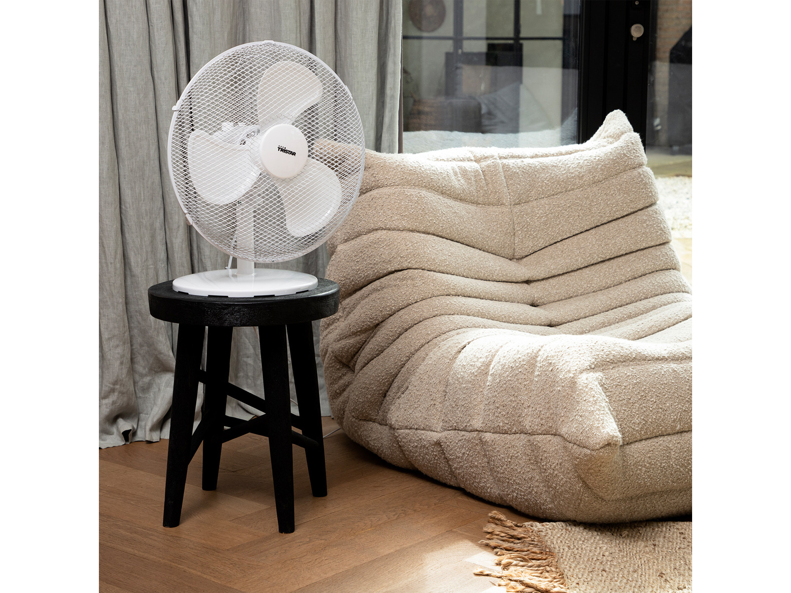 Weißer Ventilator auf einem schwarzen Hocker neben einem beigefarbenen Sitzsack und einem grauen Vorhang.