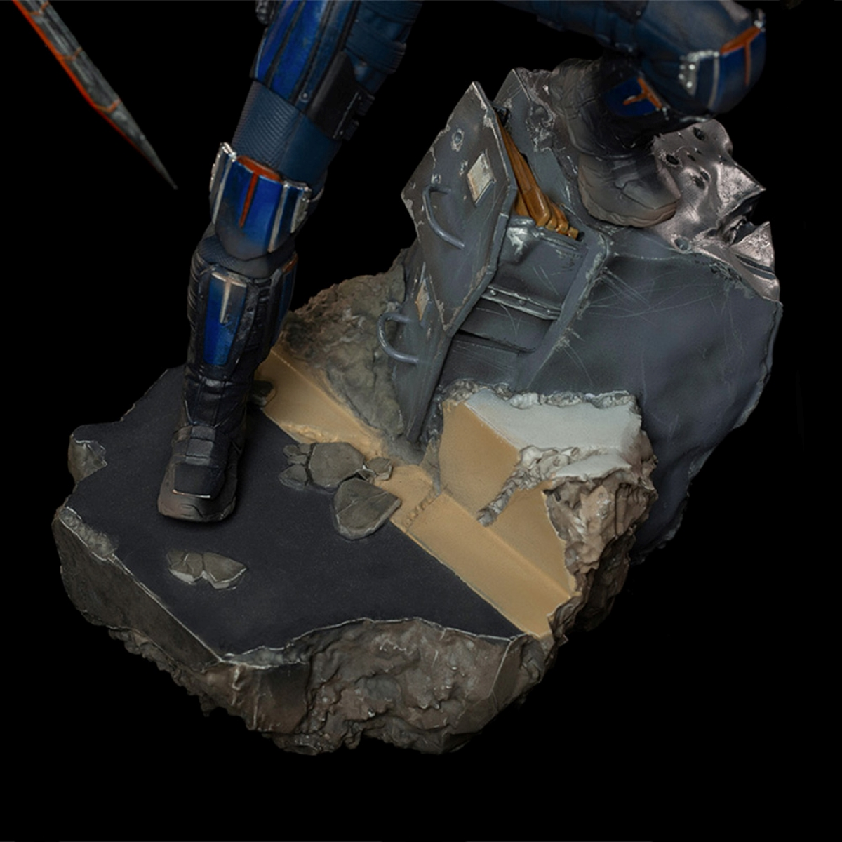 Ein Figurenfuß. Es hat einen schwarzen Schuh auf einer strukturierten, grauen Oberfläche. Der Hintergrund ist schwarz.