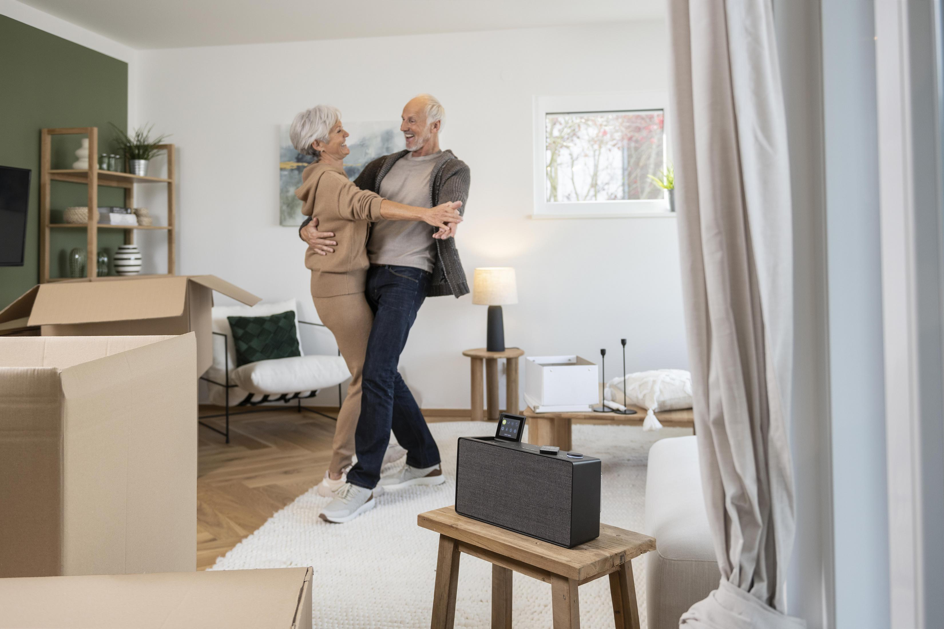 Älteres Ehepaar tanzt in einem Wohnzimmer mit Umzugskartons und einem Lautsprecher auf einem Hocker.