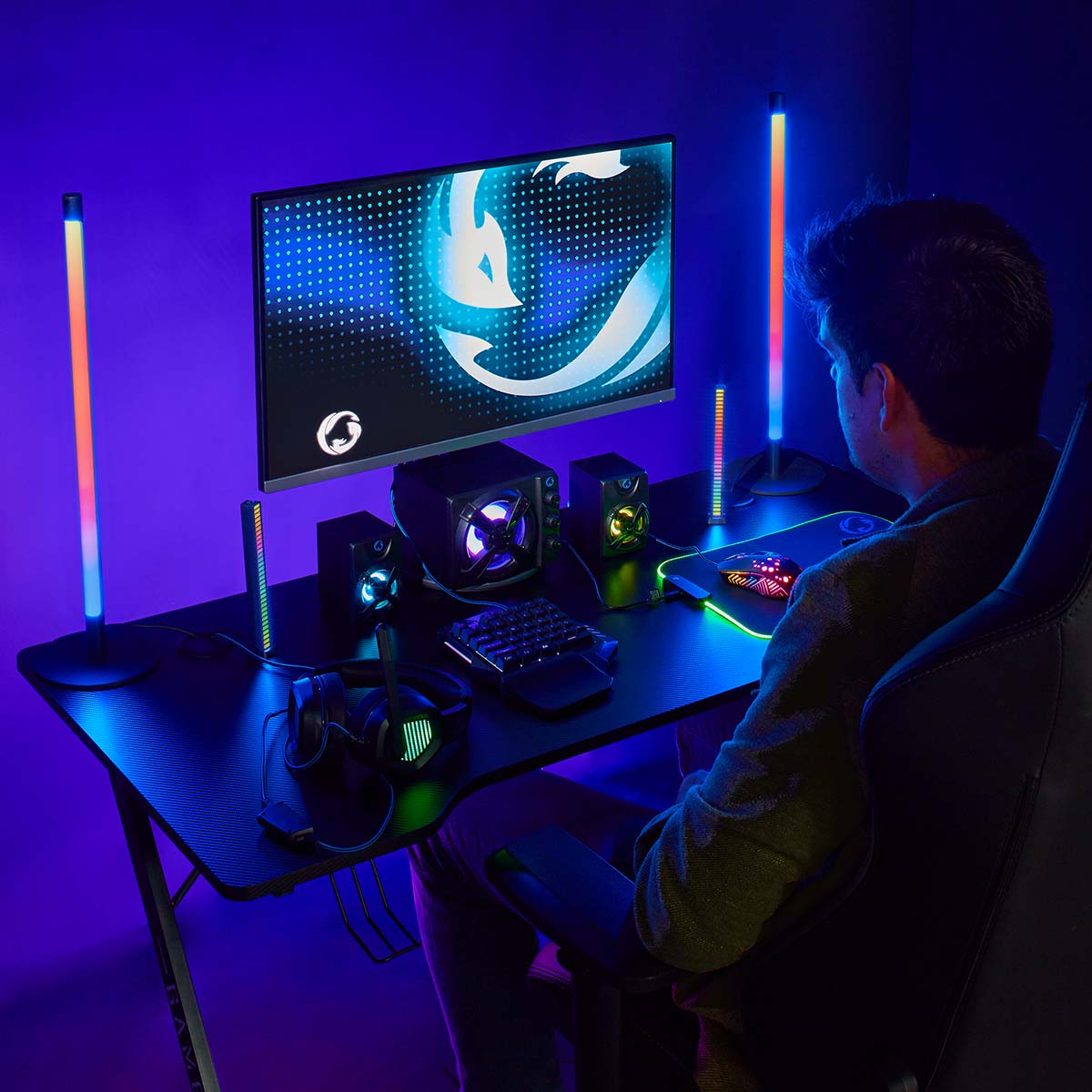 Eine Person sitzt an einem Gaming-Schreibtisch mit Monitor, Lautsprechern, Tastatur und Lichtern. Der Hintergrund ist lila.