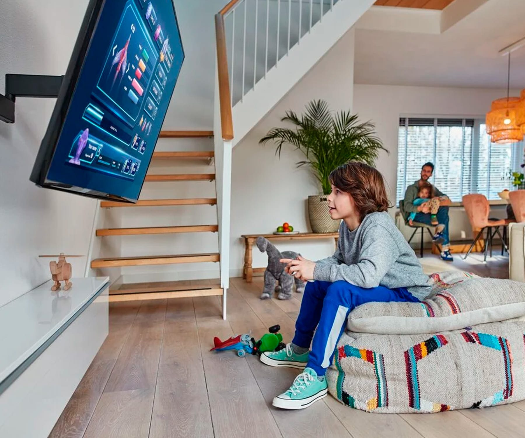 Un niño sentado mirando la televisión montada en la pared. Una escalera es visible. Familia en el fondo.
