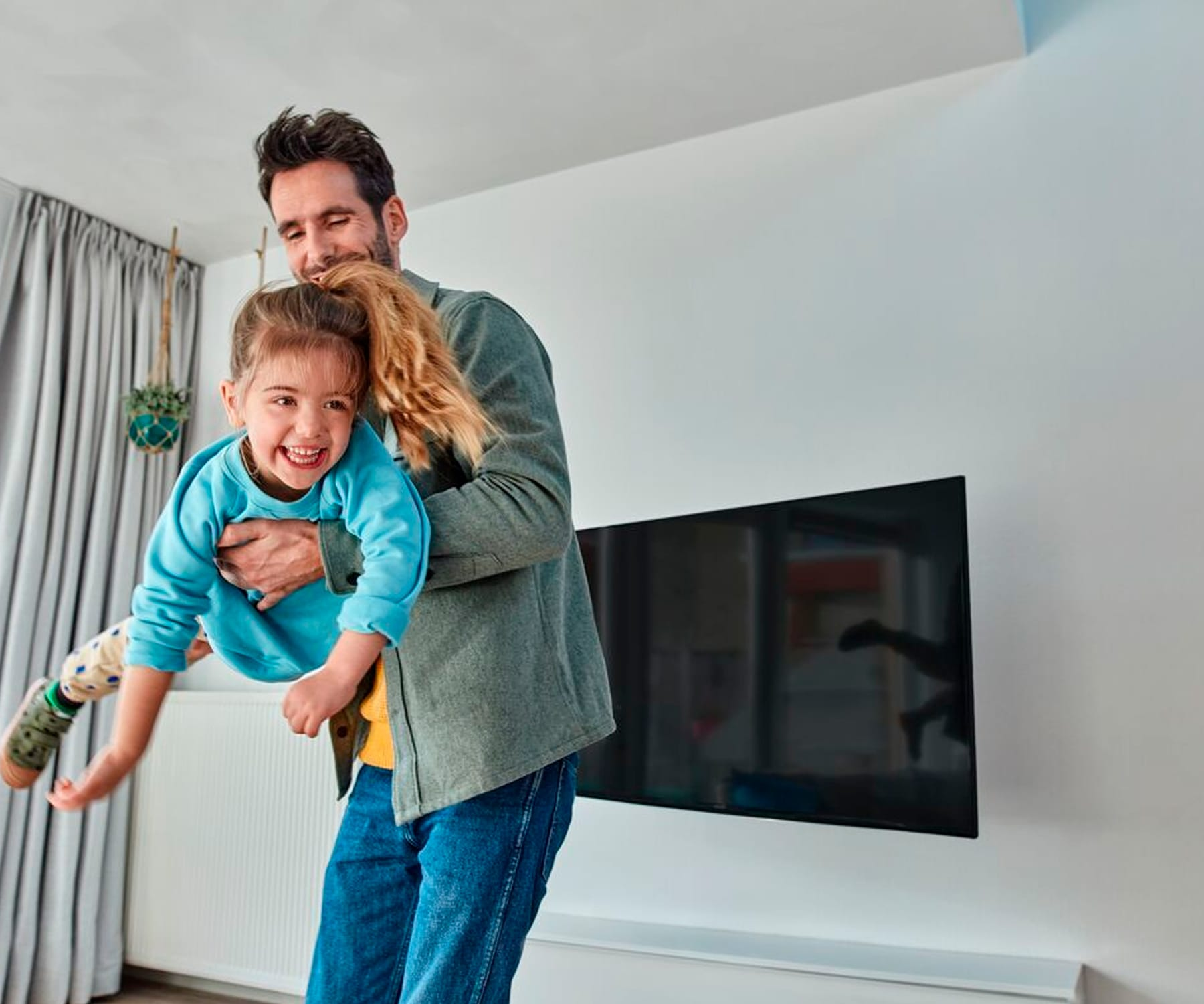 Un padre sosteniendo a su hija riendo. Están en el interior, cerca de una pared y un televisor.