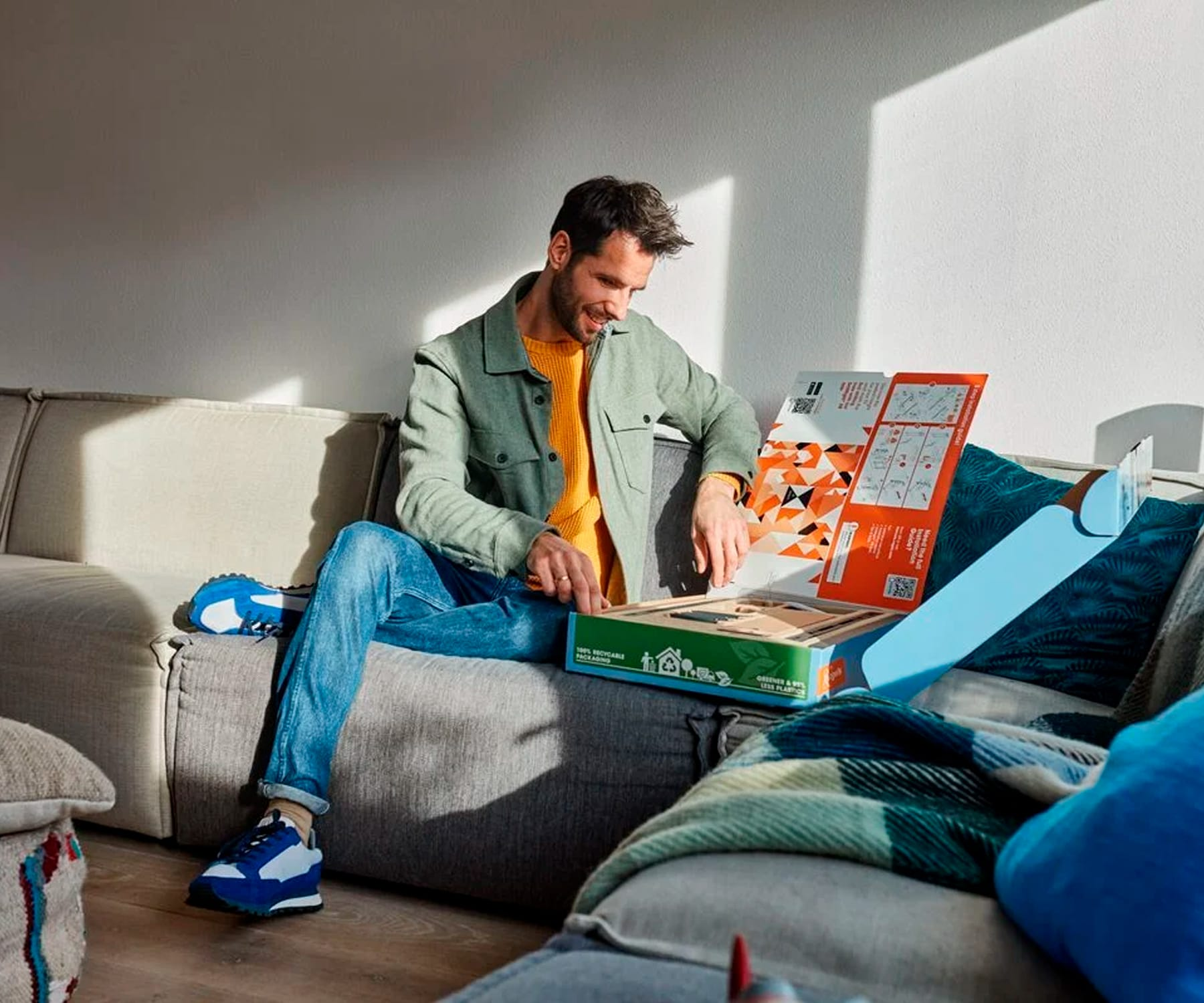 Mężczyzna siedzi na kanapie otwierając pudełko z roślinami i narzędziami. Światło słoneczne. Scena w salonie.