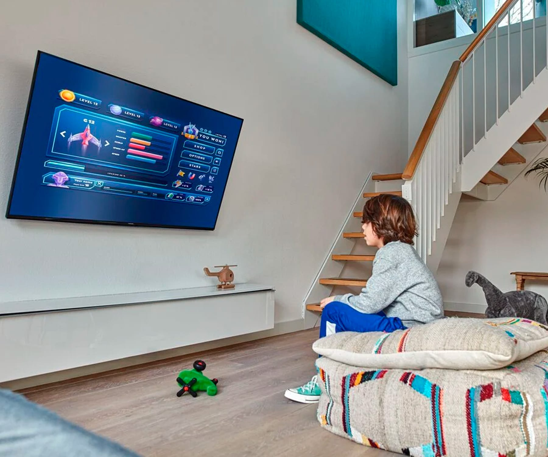 Ein Junge sitzt und sieht ein Spiel auf einem an der Wand montierten Fernseher, im Hintergrund sind Treppen zu sehen.