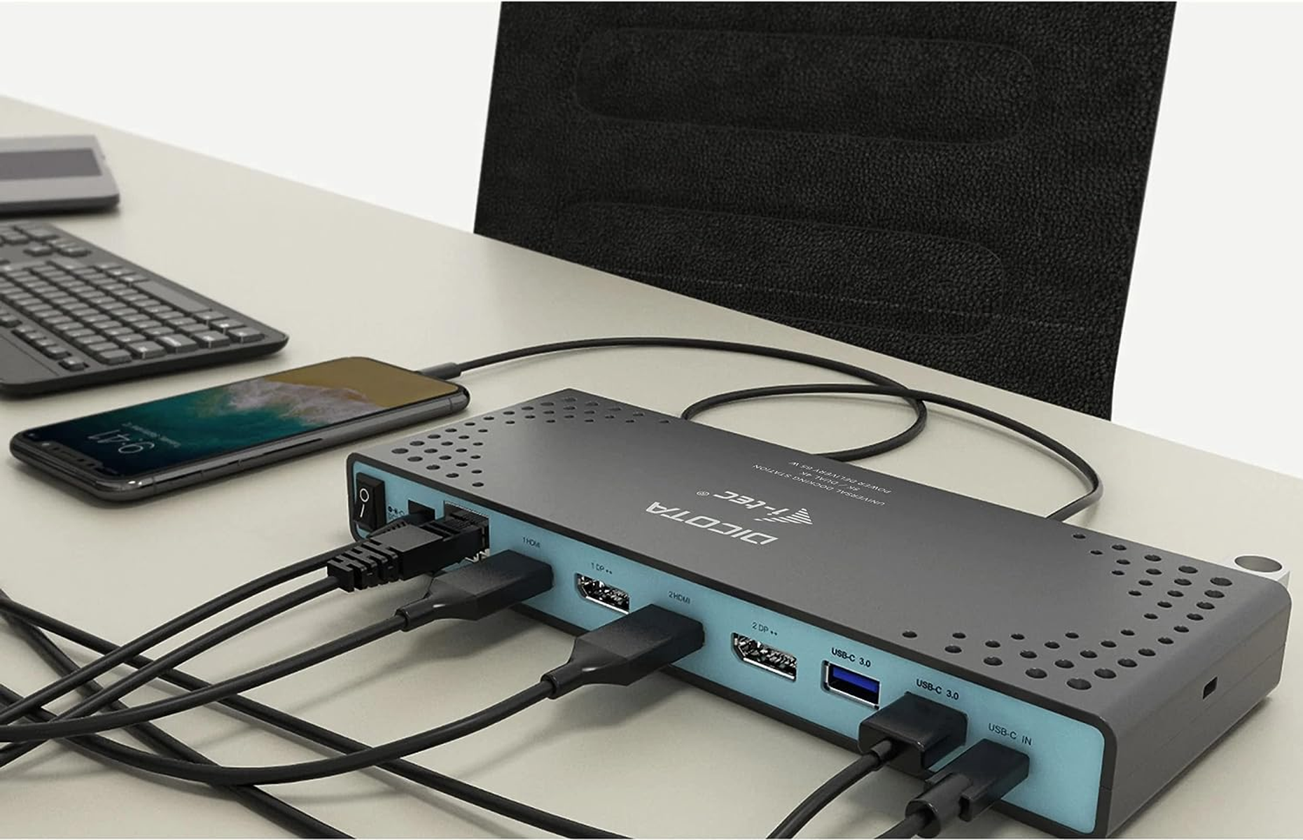 Estación de acoplamiento negra con cables conectados, un teléfono y un teclado, en un escritorio blanco.