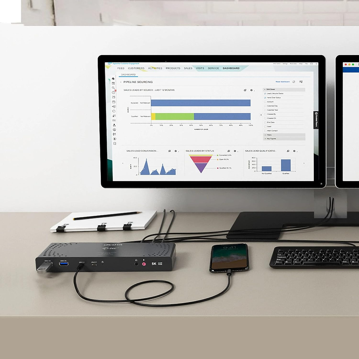 Configuración de escritorio con dos monitores mostrando datos. Un hub negro se conecta a un teléfono.