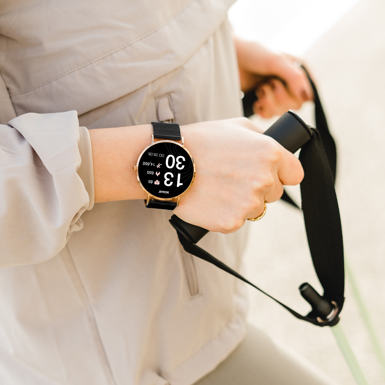 Person, die eine Smartwatch mit angezeigter Uhrzeit trägt, hält schwarze Griffe. Beige Jacke im Hintergrund.