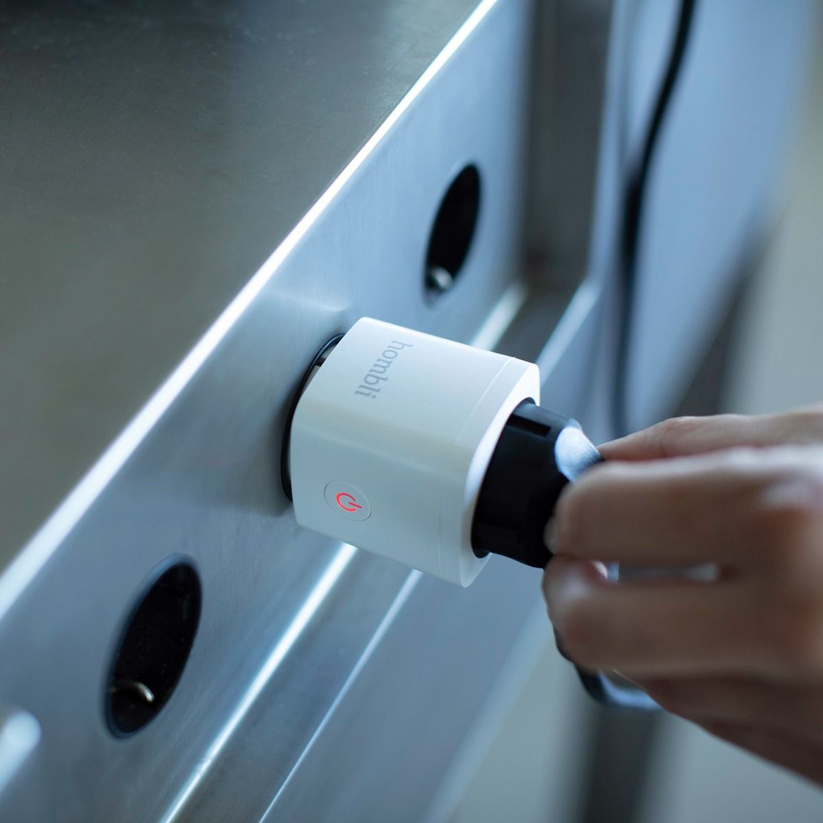 Weißer Smart Plug in der Steckdose, rotes Licht an, Hand in der Nähe. Der Markenname 'homili' ist sichtbar.