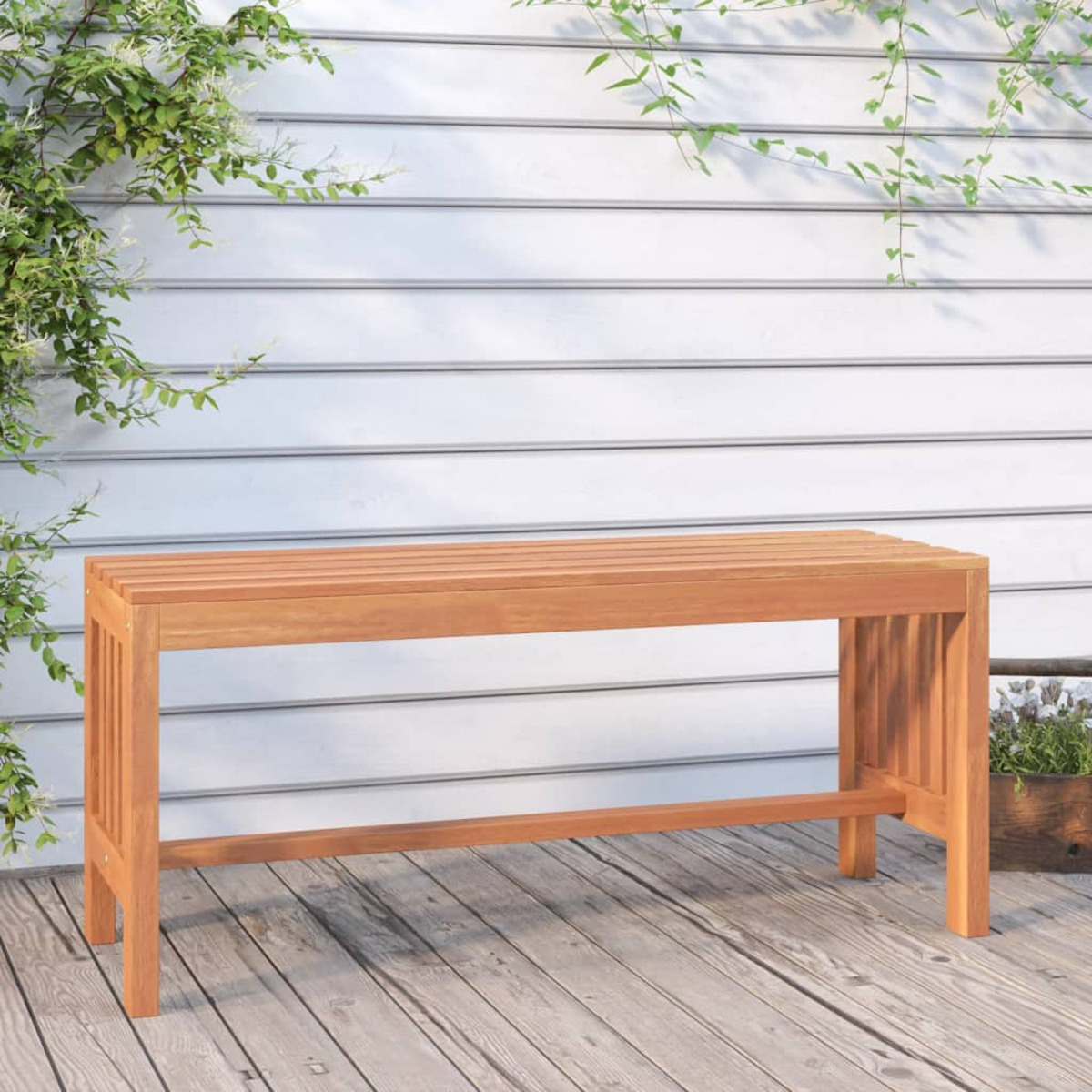 Holzbank auf einem Holzdeck, mit einer weißen Wand und grünen Pflanzen im Hintergrund.