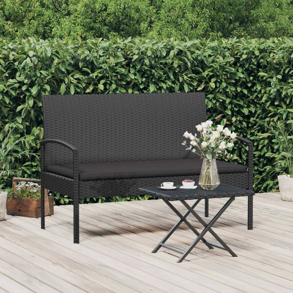 Ein schwarzes Korbbank- und Tischset auf einem Holzdeck. Im Hintergrund sind Blumen und grüne Büsche zu sehen.