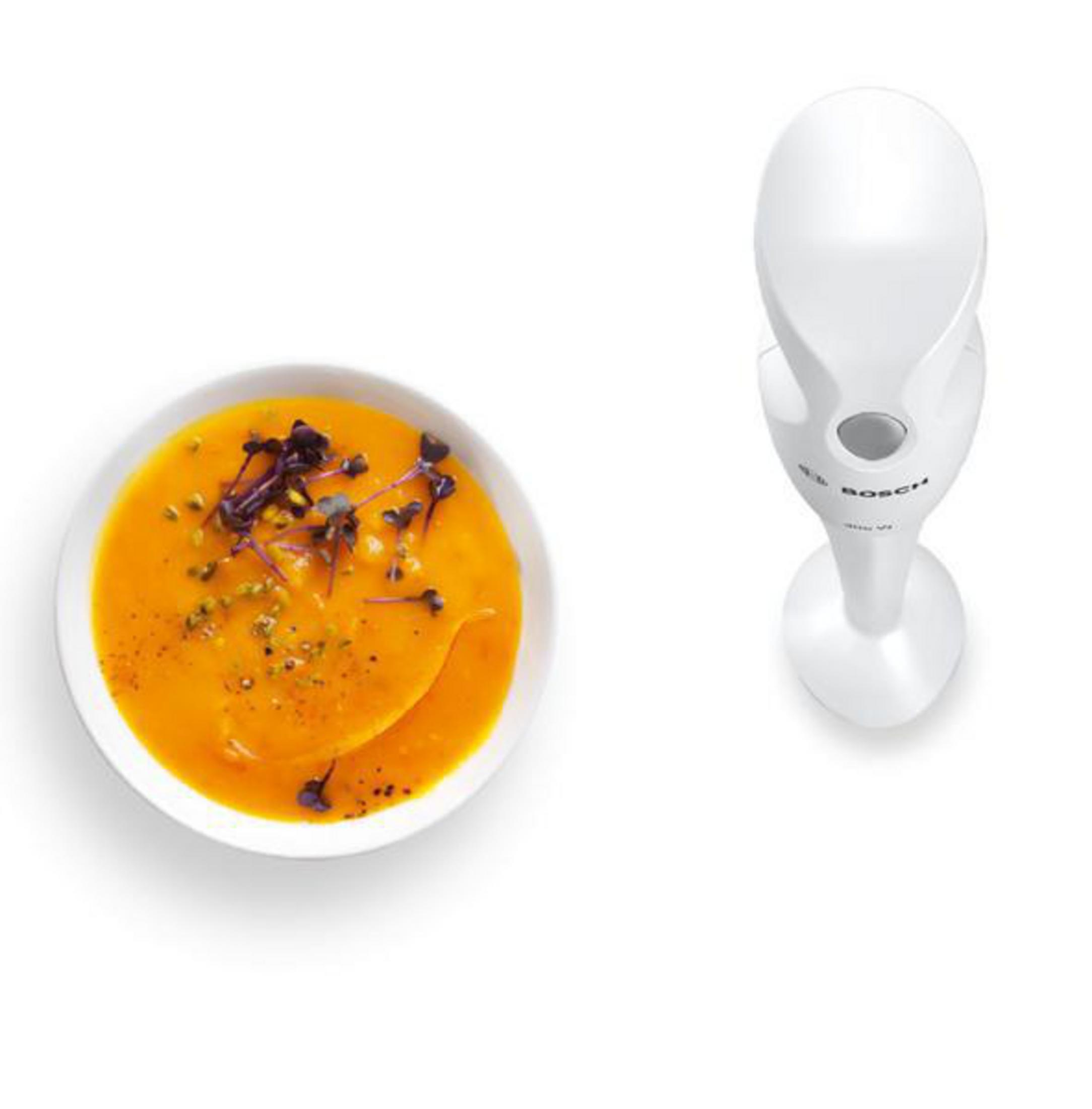 Eine Schüssel mit orangener Suppe mit Garnitur und ein weißer Stabmixer auf weißem Hintergrund.
