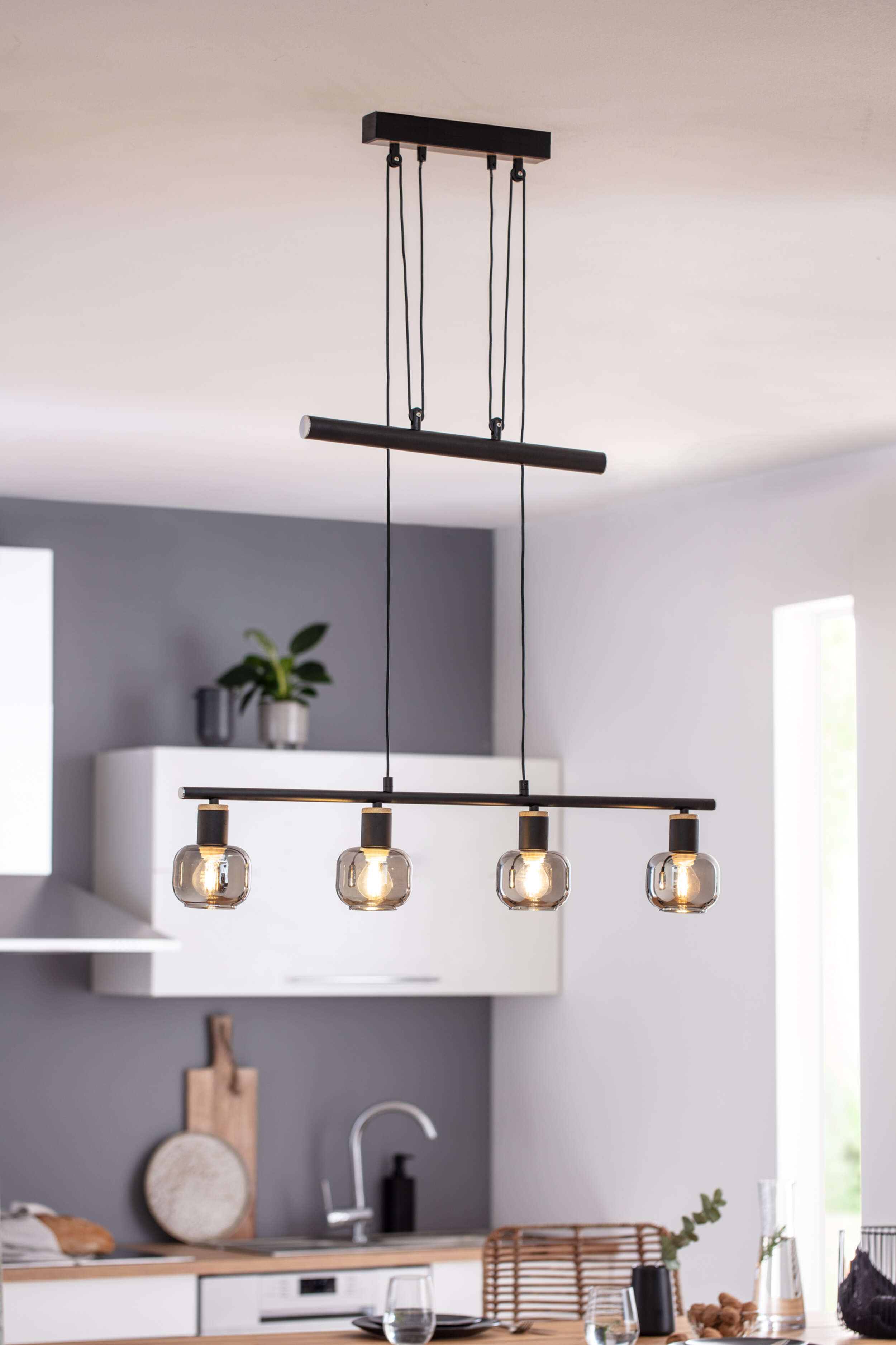 Una moderna lampada a sospensione lineare nera con quattro paralumi in vetro trasparente, in un ambiente cucina.
