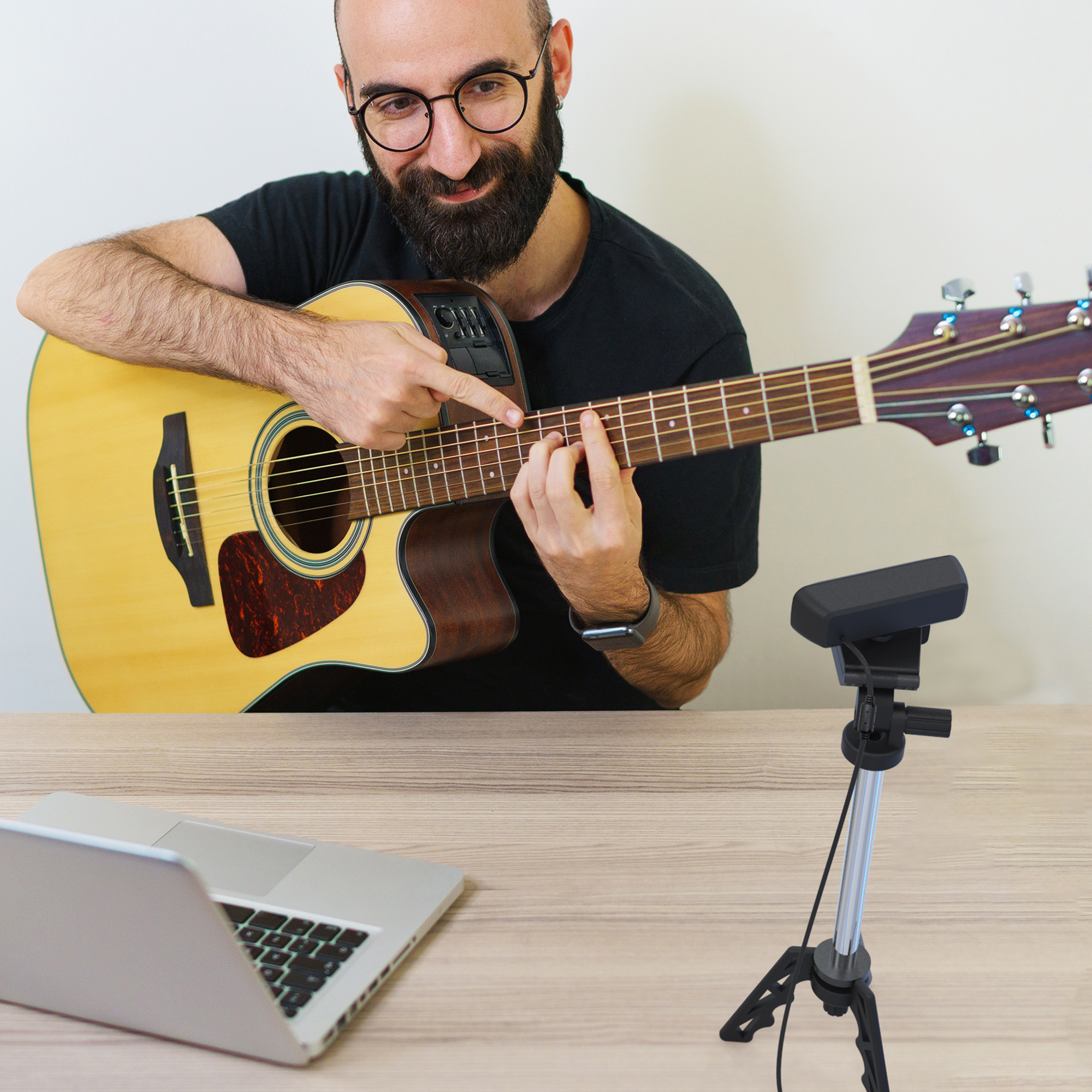 Mężczyzna gra na gitarze, wskazując na gryf, z laptopem i mikrofonem na stole.