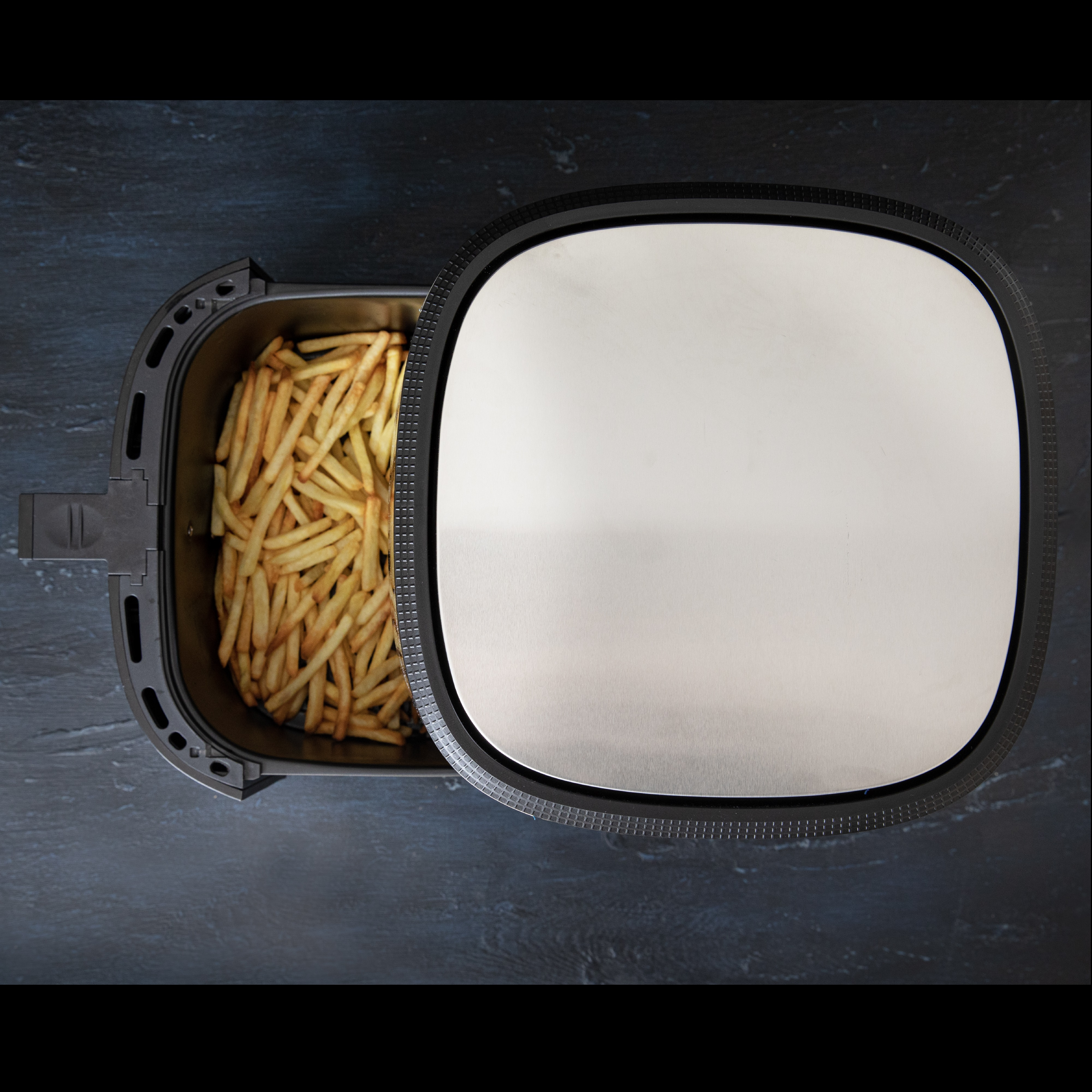 Eine offene Heißluftfritteuse mit Pommes Frites darin, der Deckel ist daneben, Draufsicht.