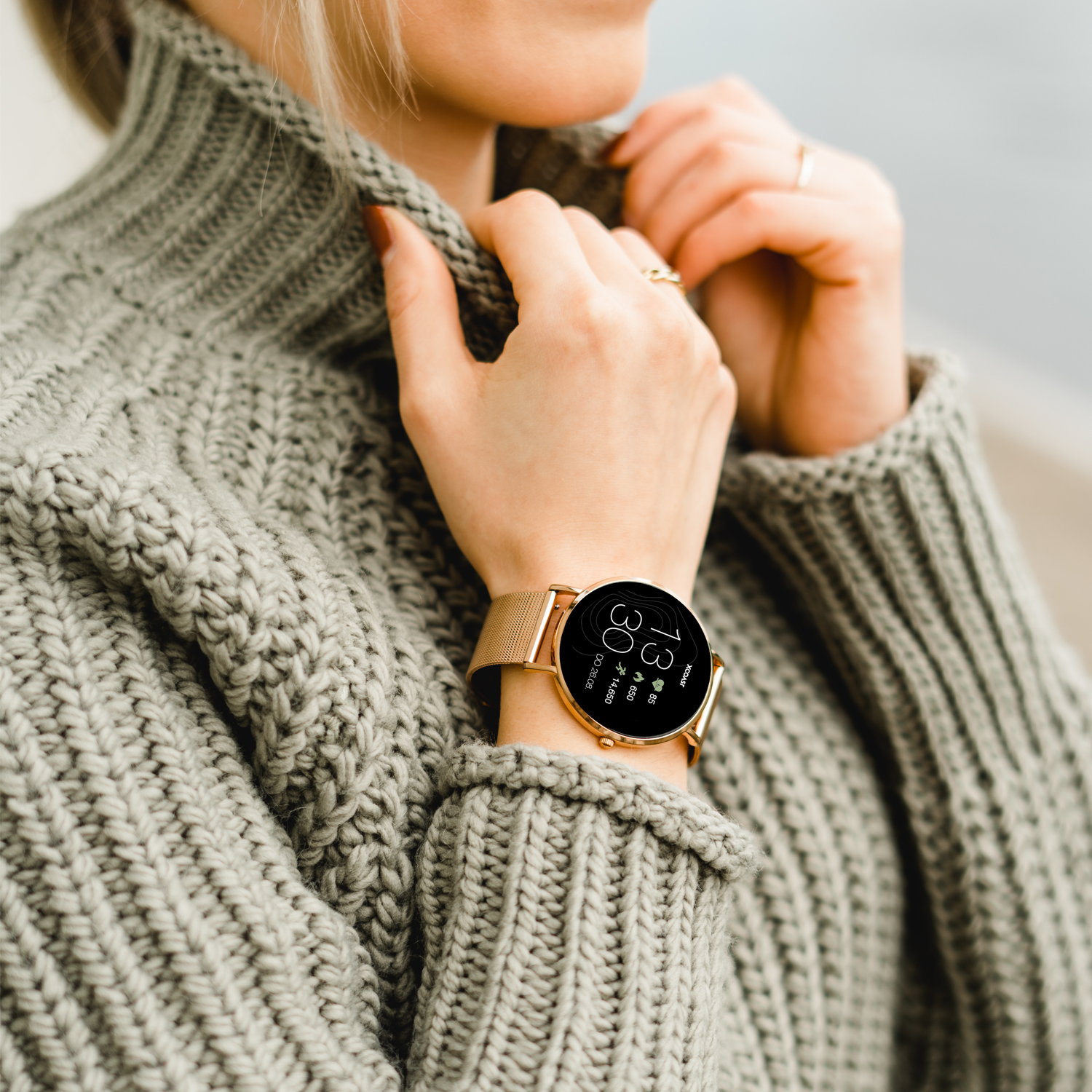 Eine Frau mit grauem Pullover und goldener Uhr am Handgelenk, verschwommener Hintergrund.