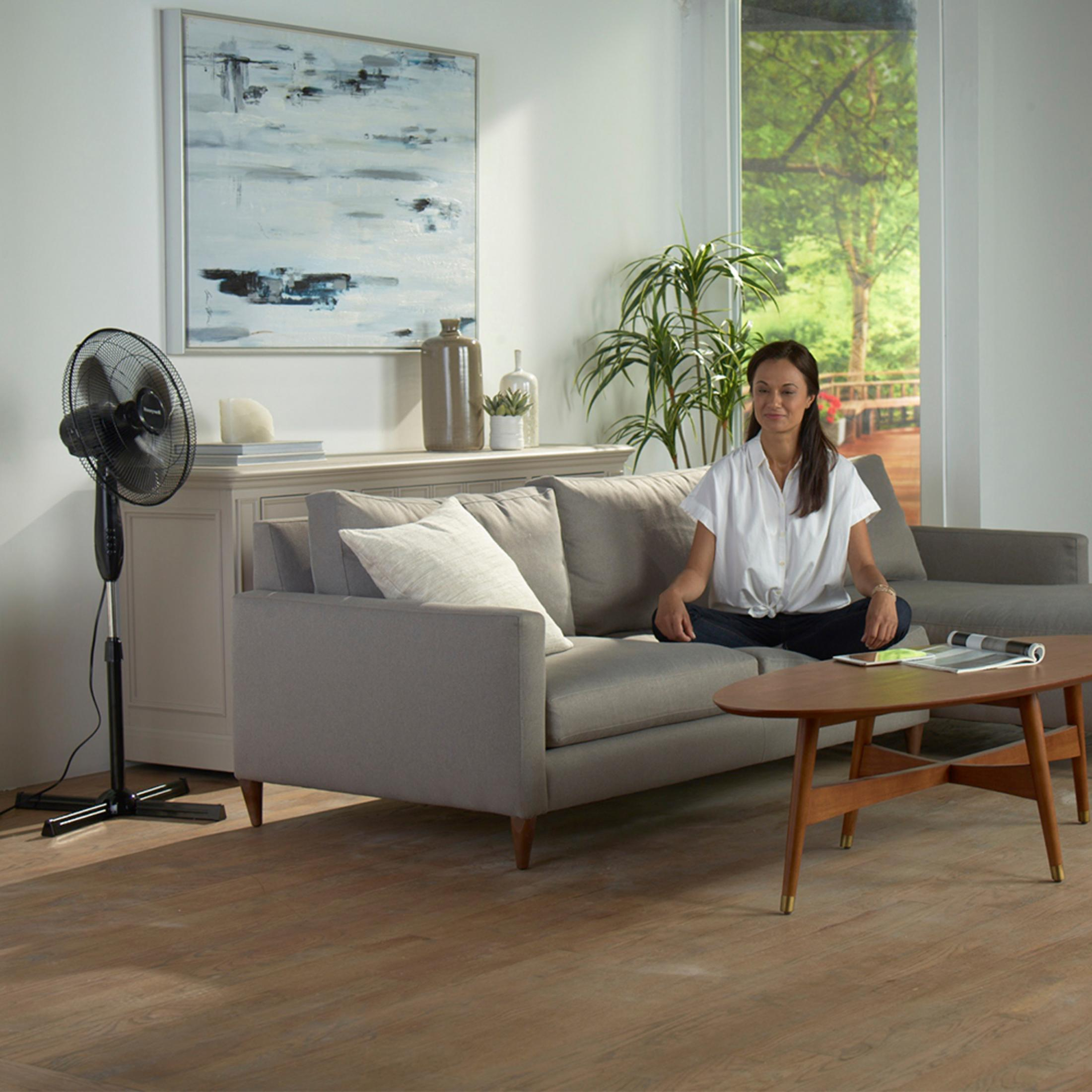Mujer sentada cerca de un sofá y un ventilador. Mesa de madera y pintura en la pared.