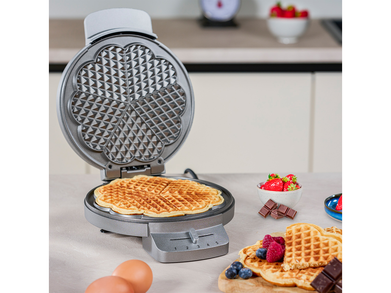 Waffeleisen offen, mit fertiger Waffel darin. Beeren und Schokolade in der Nähe.