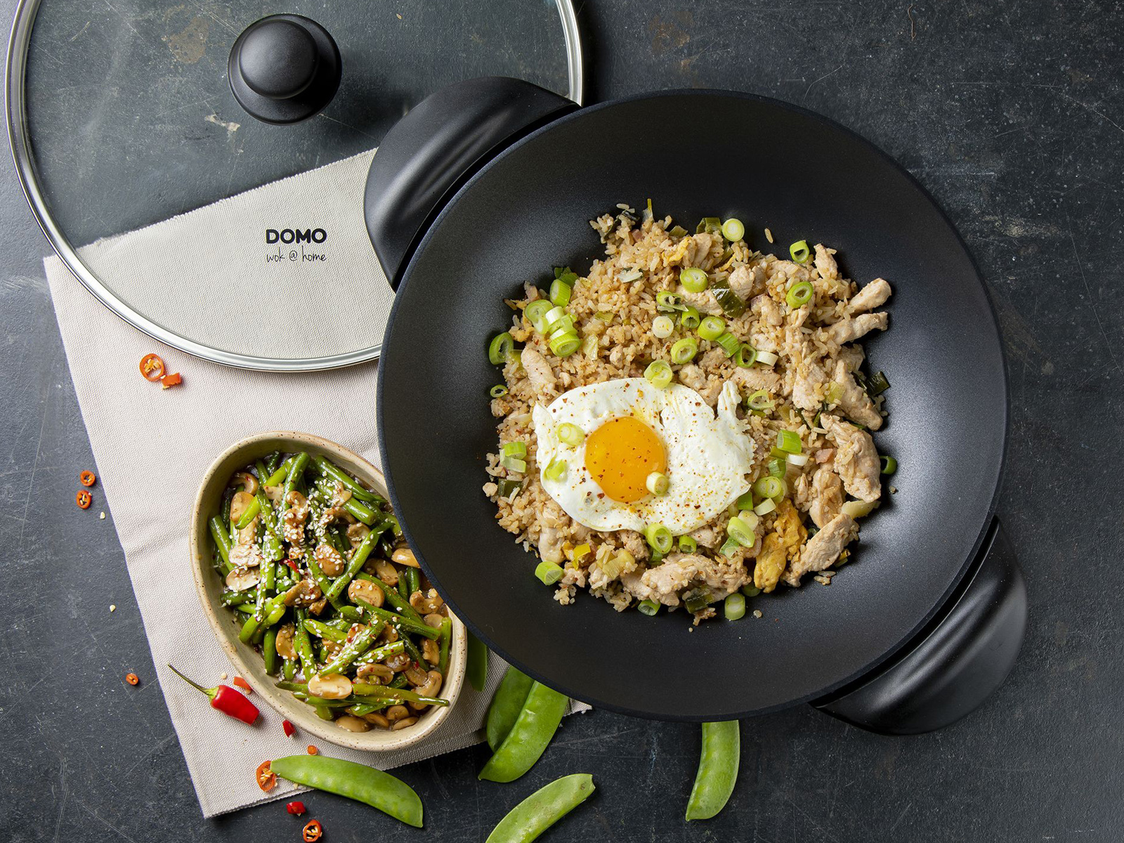 Ein Wok mit gebratenem Reis, Ei und Frühlingszwiebeln. Außerdem eine Beilage und ein Wokdeckel.