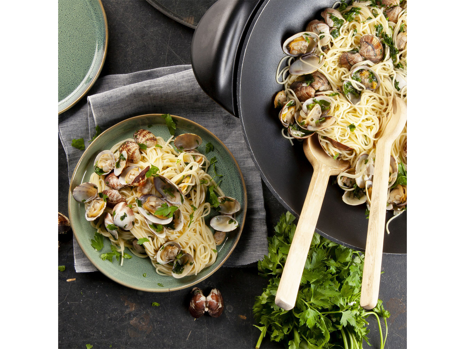 Pasta mit Muscheln auf einem Teller und in einer Pfanne serviert, mit Holzlöffeln und Petersilie.