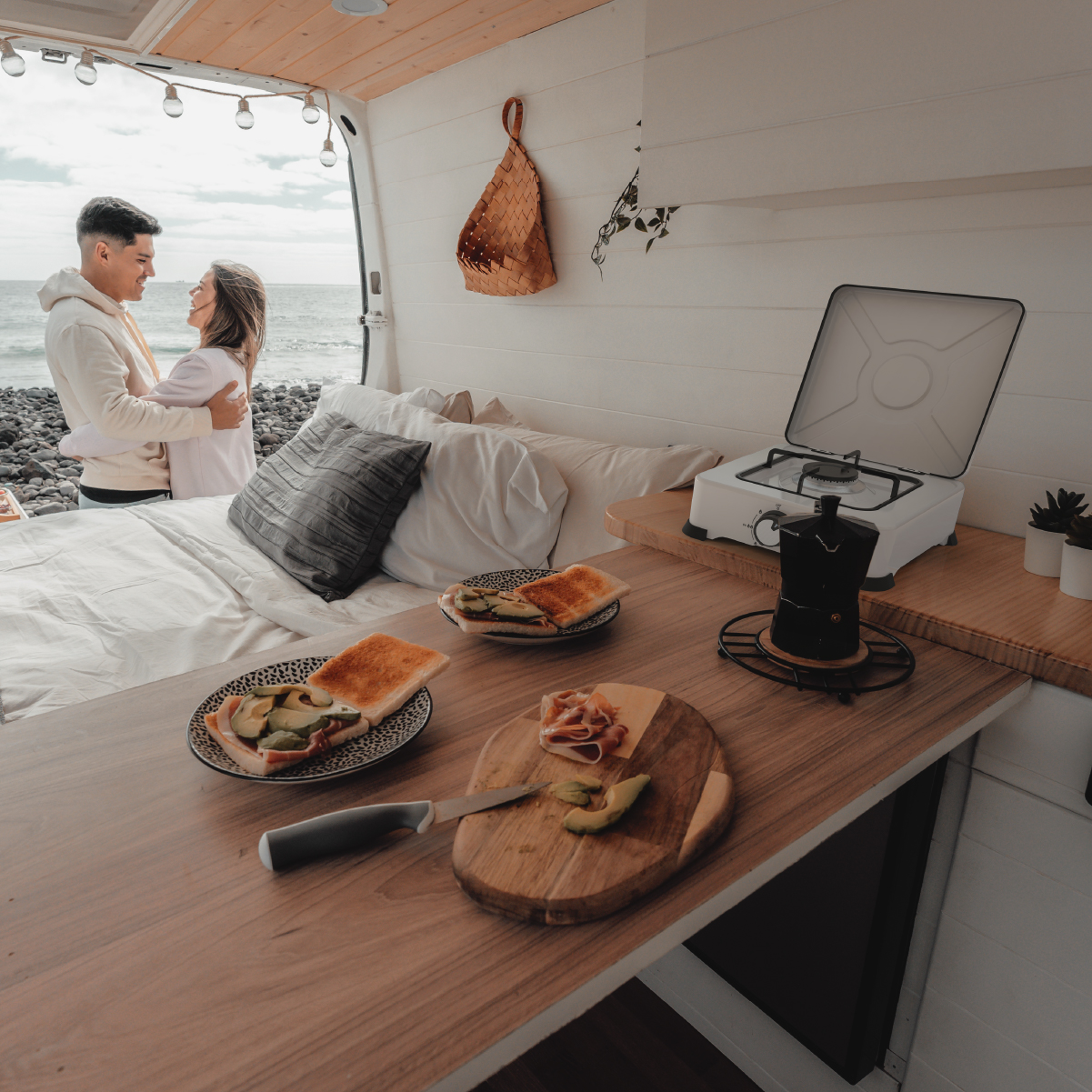 Una coppia si abbraccia in un camper vicino al mare. Sulla tavola ci sono panini e un fornello.