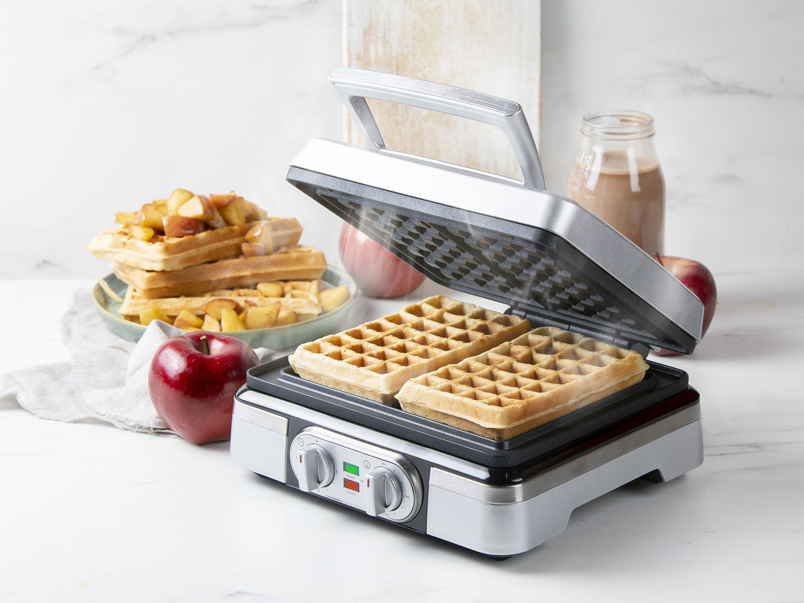 Ein Waffeleisen mit zwei Waffeln. Äpfel und eine Milchflasche sind ebenfalls vorhanden.