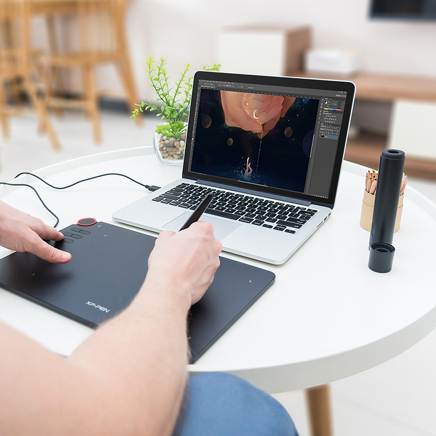 Hände benutzen Stift und Tablet, verbunden mit Laptop mit Kunstwerk. Kleine Pflanze steht daneben.