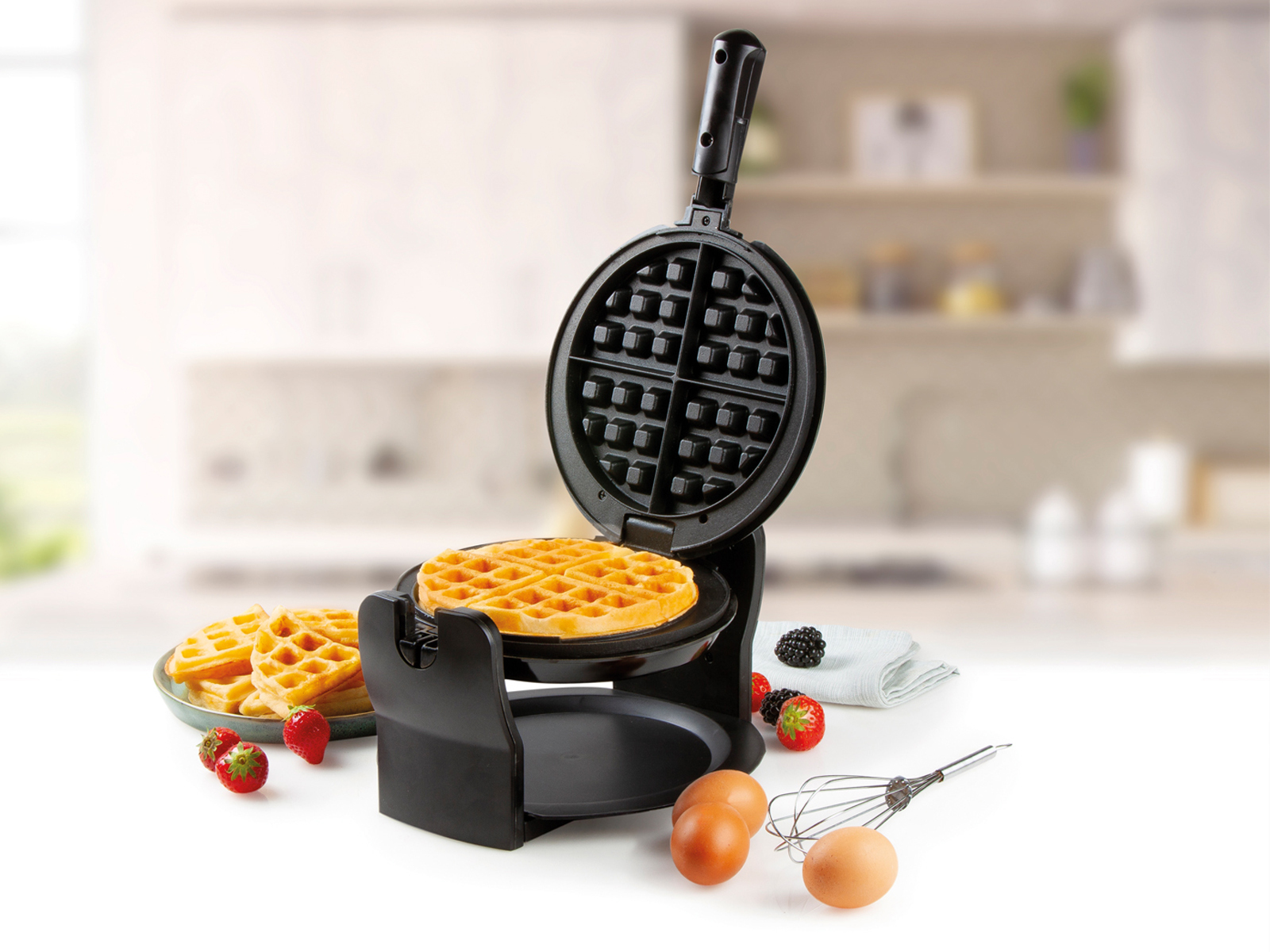 Waffeleisen mit Waffel, Waffeln, Eiern und Früchten auf weißer Oberfläche.