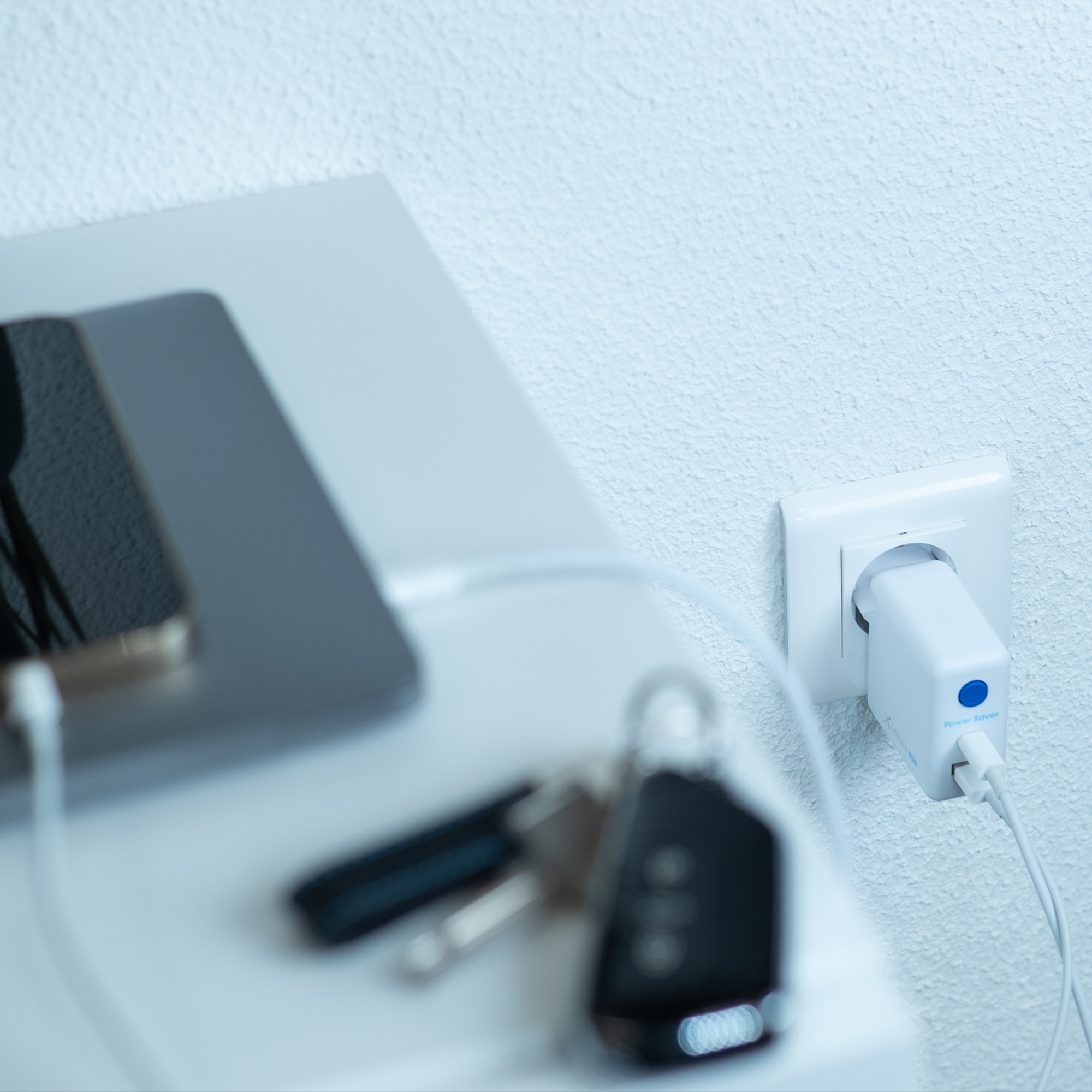 Weißer Nachttisch mit Laptop, Schlüsseln und ladendem Telefon. Netzteil in der Wand.