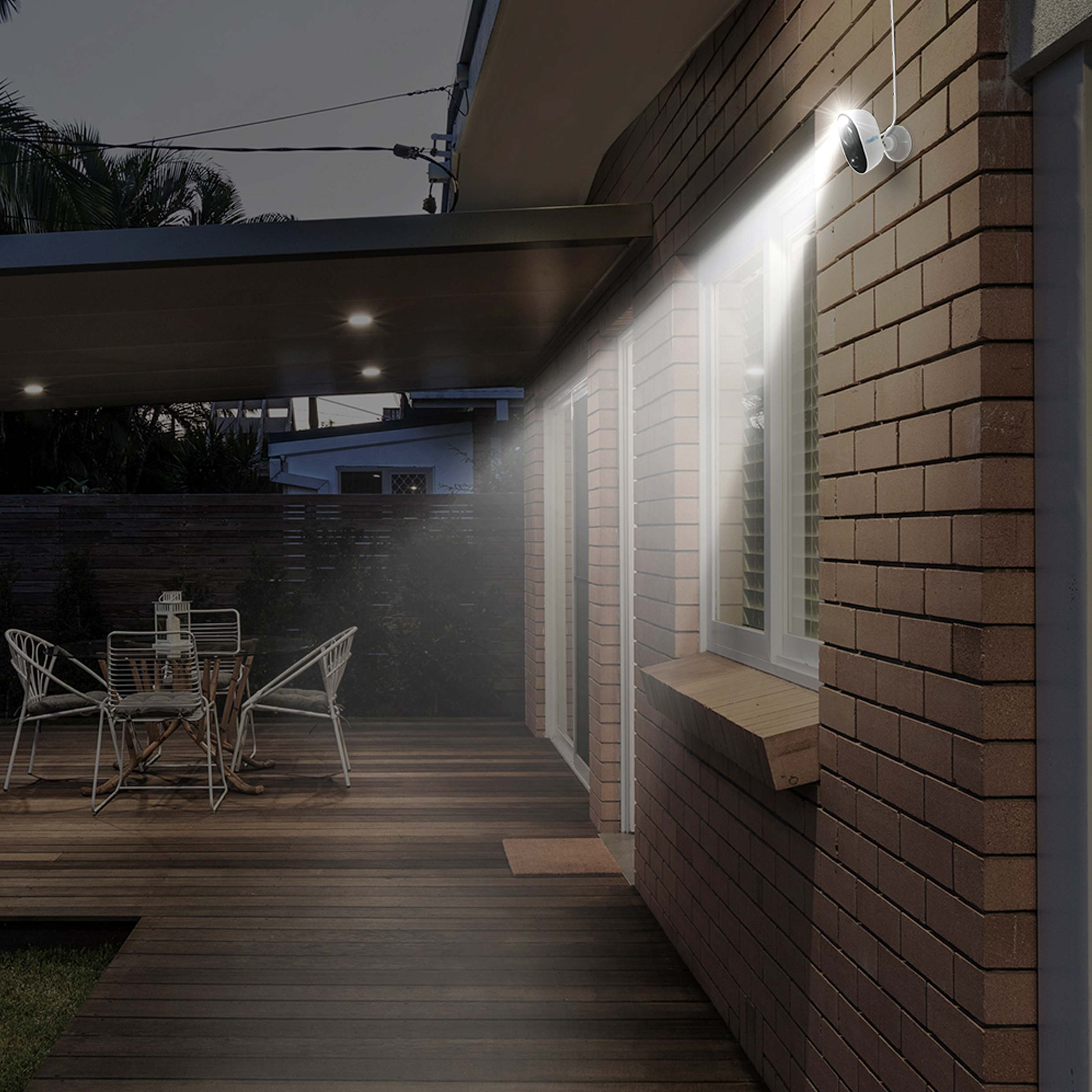 Scena zewnętrzna nocą. Ściana z reflektorem i drzwiami. Ciemny drewniany taras i krzesła.