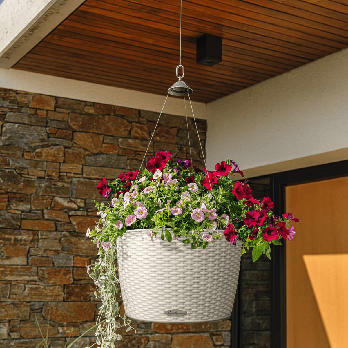 Hängender weißer Korb mit roten und rosa Blumen. Steinmauer und Holzdecke im Hintergrund.