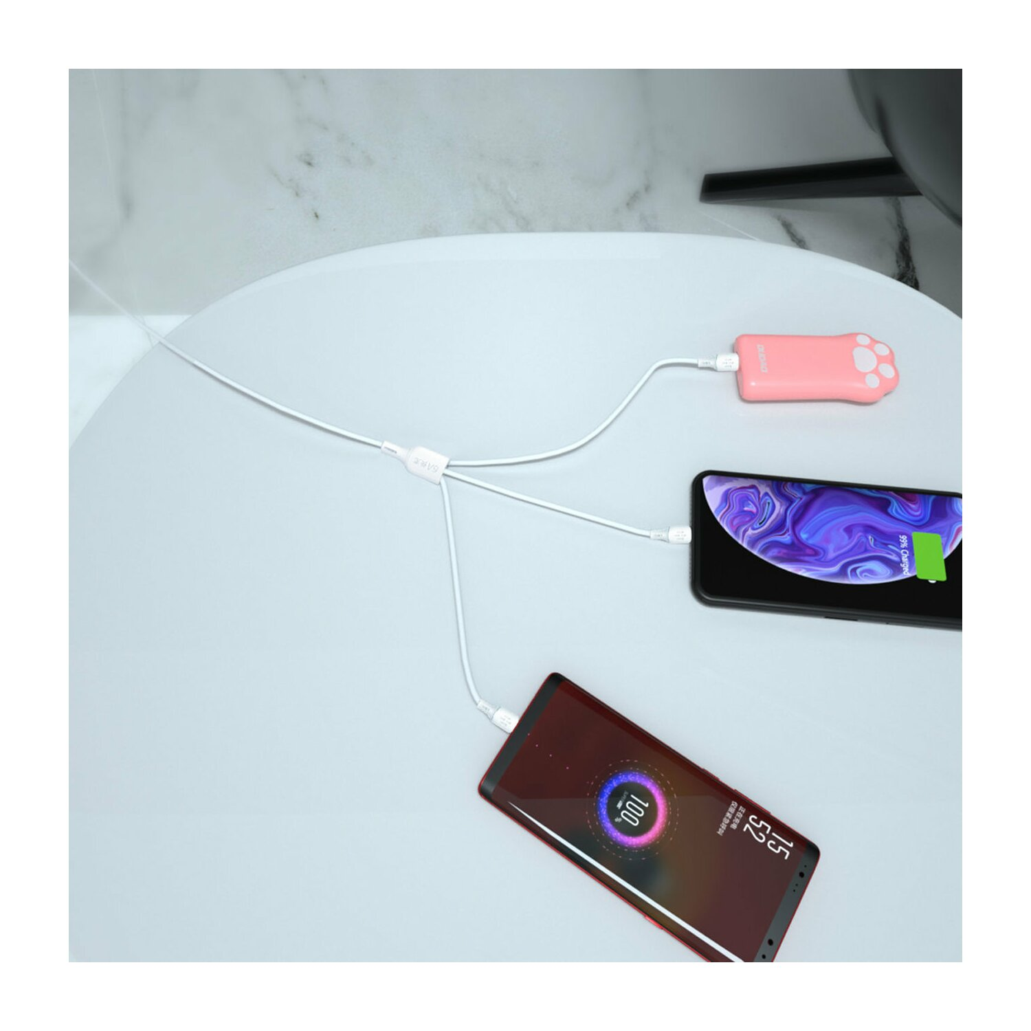 Trzy telefony ładujące się na białym stole. Jeden jest różowy, dwa czarne. Widoczna różowa ładowarka.