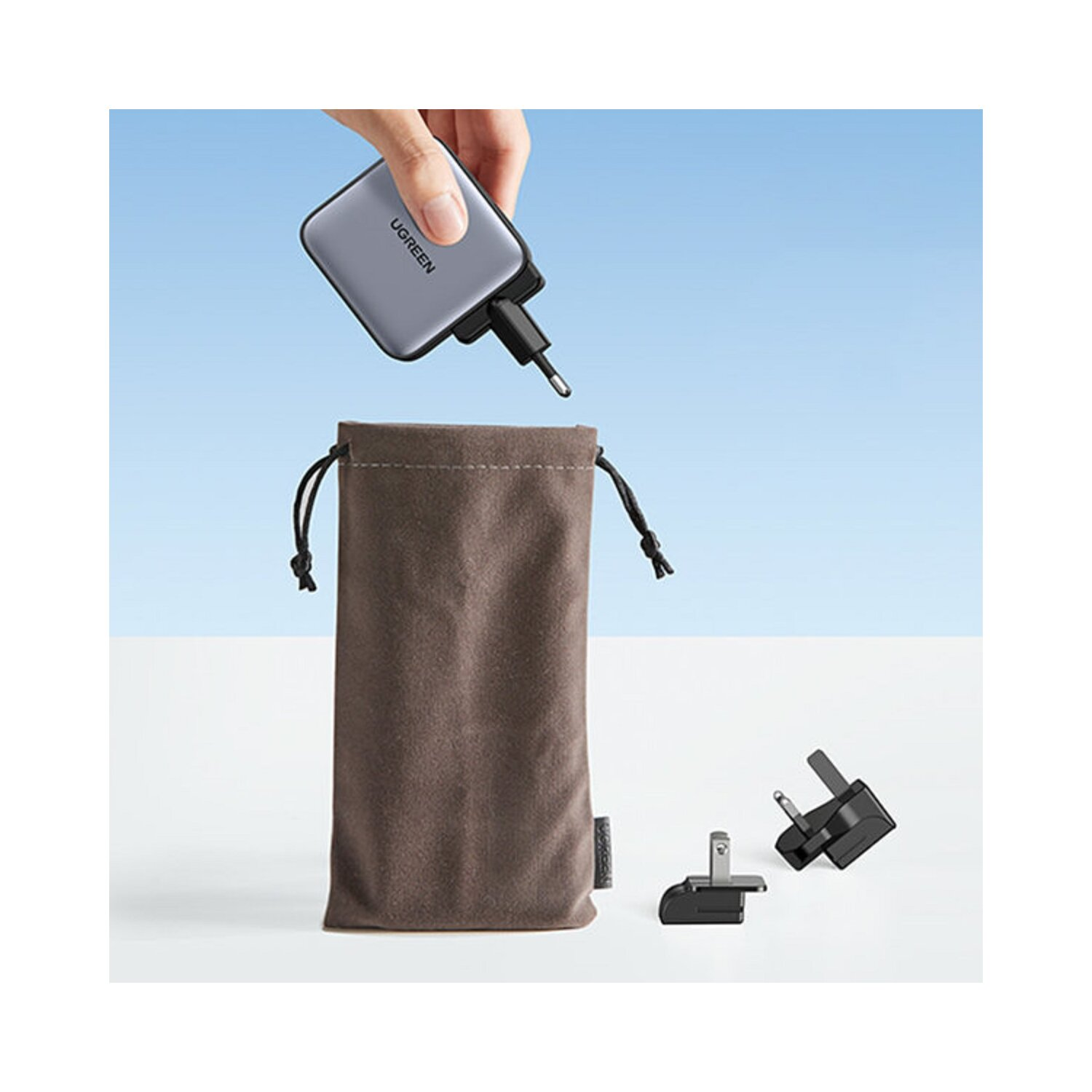 Osoba trzymająca szarą ładowarkę i brązową torbę. Białe tło.