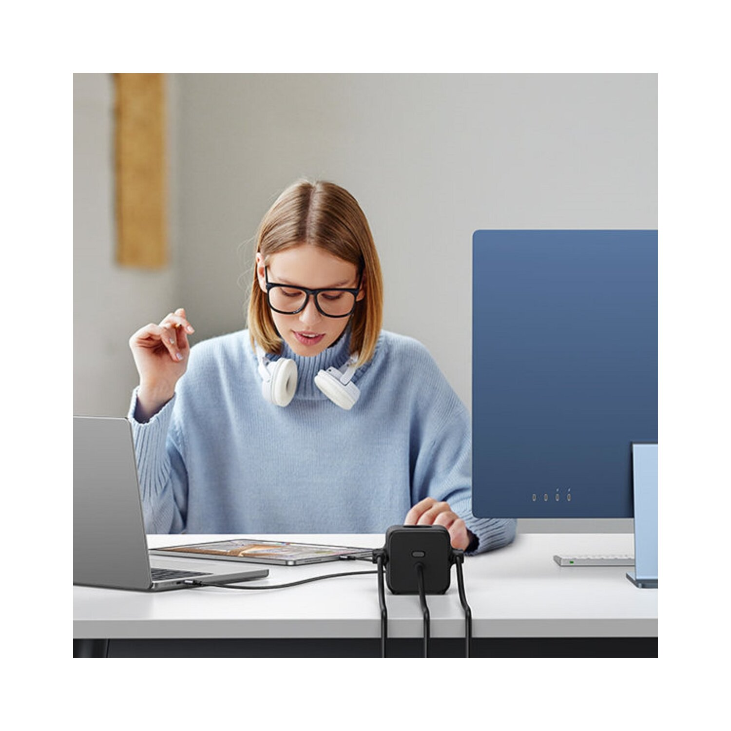 Kobieta w okularach, używająca laptopa, z zestawem słuchawkowym i niebieskim monitorem.