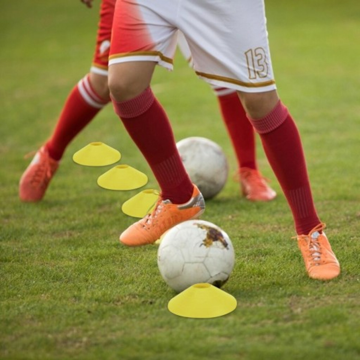 Fußballspieler in roten Socken und orangen Schuhen. Gelbe Hütchen und Fußbälle auf dem grünen Feld.